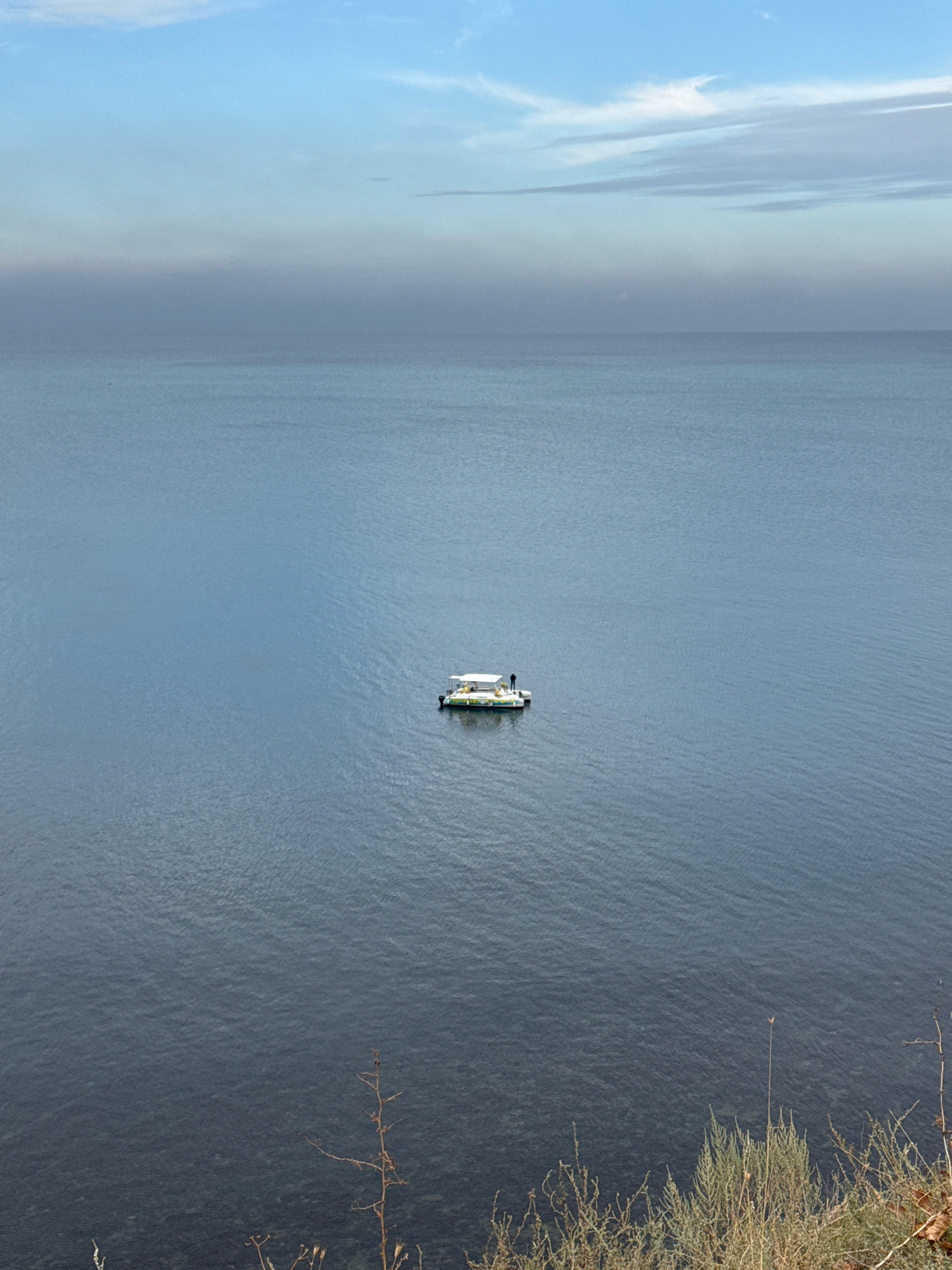 A low-angle, wide shot captures a calm, expansive sea under a hazy sky. A small, white boat with a green trim is positioned in the middle of the frame, afloat on the tranquil water. A lone figure stands on the deck of the boat, facing away from the camera. The foreground features dry, sparse vegetation, characteristic of Anapa's coastal landscape. The scene evokes a sense of quiet solitude.