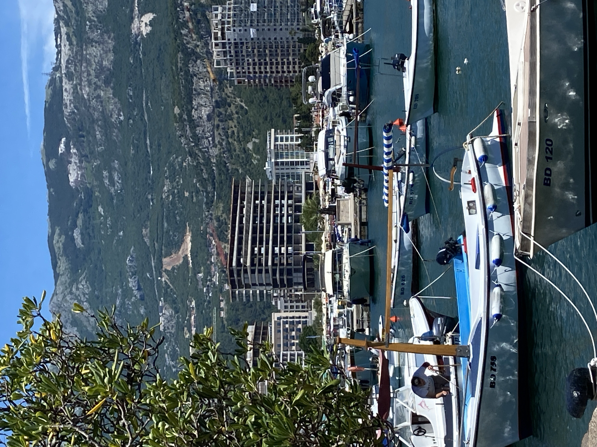 The image shows a view of the marina in Budva, Montenegro, with several boats docked in the clear blue water. In the foreground, a white boat with blue trim is visible, displaying the registration number "BD 758" on its side. Another boat, also white with blue and red trim, is partially visible in the lower right corner, marked "BD 120." Further back, a person is seen working on a white boat. In the middle ground, several larger yachts and motorboats are moored. Behind the marina, a modern city skyline rises with several multi-story buildings, some appearing to be under construction, evidenced by scaffolding and exposed frameworks. Beyond the city, lush green mountains with rocky outcrops form the backdrop. The sky is a clear, bright blue, indicating fair weather. A tree with green leaves partially obstructs the upper left portion of the scene.