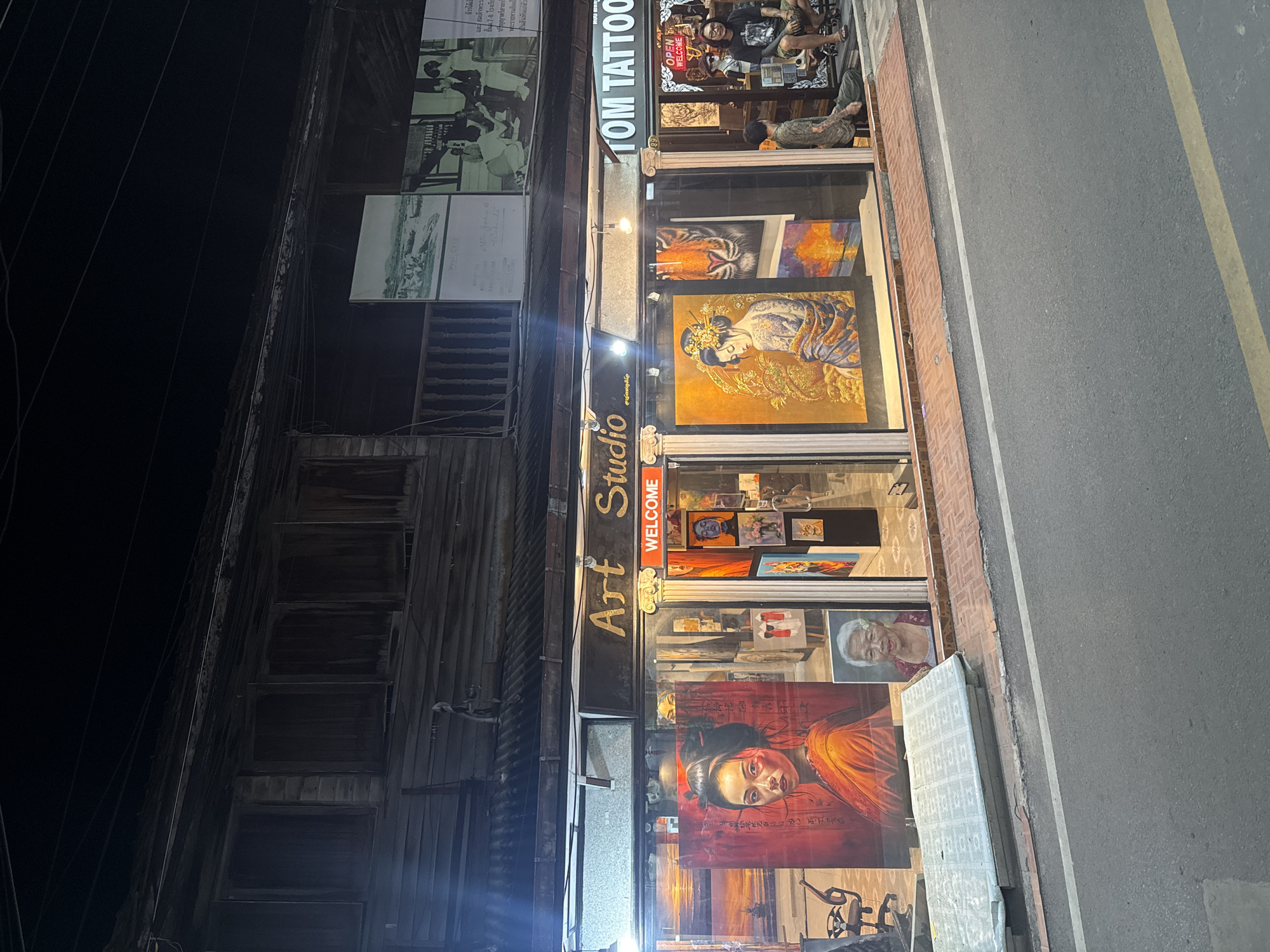 The image captures a street-level view of a shopfront in Hua Hin City Municipality, Thailand, at night. The shop appears to be an art studio and tattoo parlor, with a prominent "Art Studio" sign above the entrance. Large paintings are displayed in the window, including a portrait of a woman in traditional attire, a tiger's face, and a geisha. To the right, the "TOM TATTOO" sign is visible, with an "OPEN" sign in the window and two individuals sitting outside the shop. The building has a traditional wooden facade with multiple windows. A street with a white line and a yellow stripe runs in front of the shop, with a sidewalk made of red bricks. A mattress is stacked on the sidewalk. Overhead, power lines crisscross the dark sky. The lighting suggests it is nighttime.