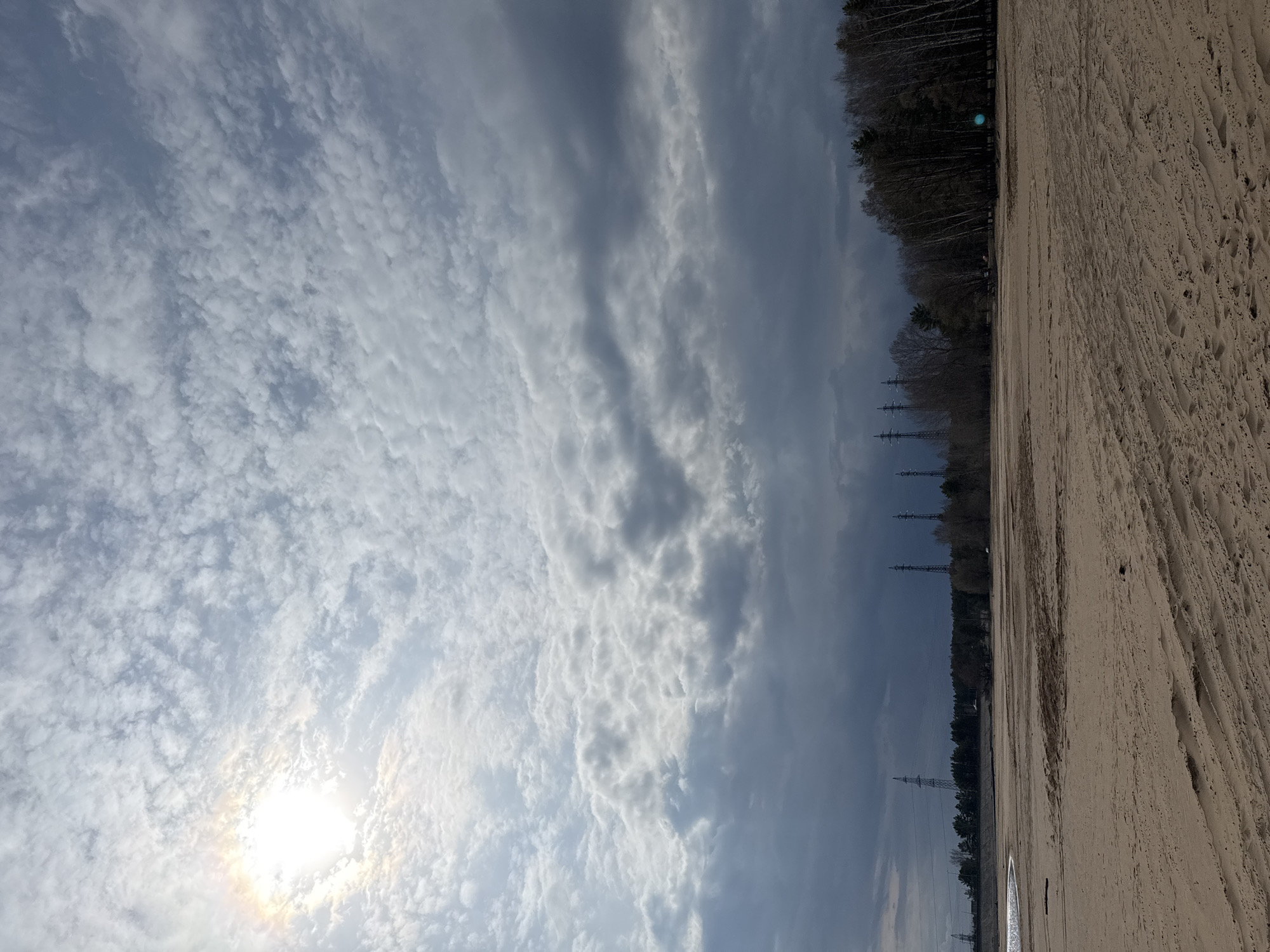 The image depicts a landscape scene in Noviy, Russia. The ground appears to be a sandy area, with visible tire tracks. A line of trees marks a boundary along the horizon. Beyond the trees, power lines are visible against a cloudy sky. The sky itself is a mix of light blue and gray, with patches of dense clouds and a bright sun partially obscured.