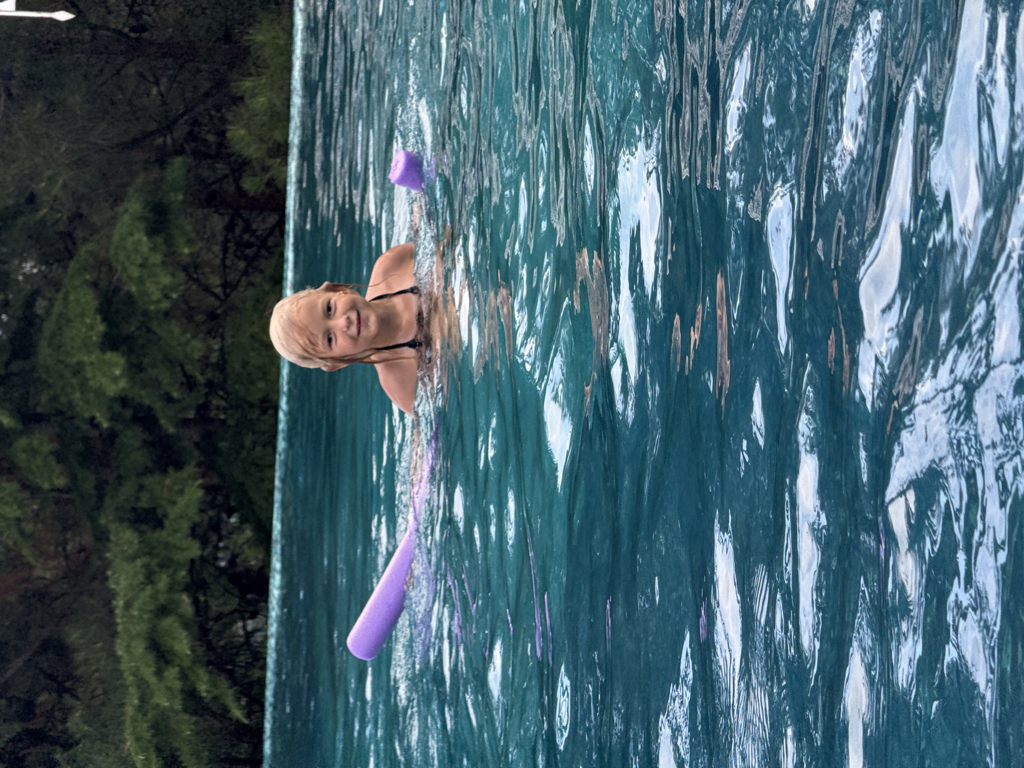 In the city of Yalta, Ukraine, a young girl with blonde hair is swimming in a pool, holding a purple pool noodle. She is smiling at the camera. The water is a vibrant blue-green, with ripples on the surface. Behind her, lush green trees line the edge of the pool.