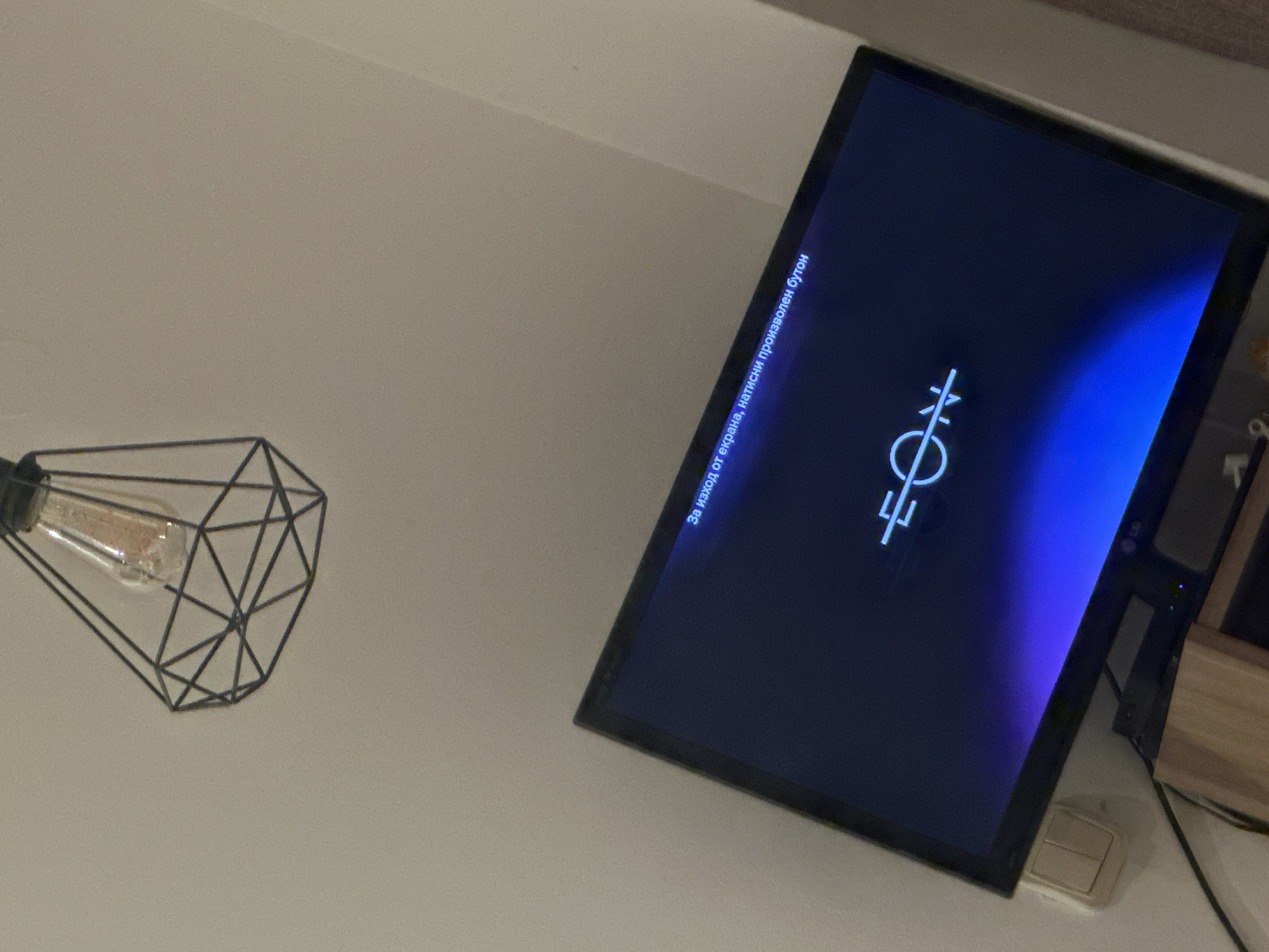 The image shows a close-up of a black-framed LG computer monitor displaying a symbol consisting of a circle with two intersecting lines through its upper half, topped by two short vertical lines. A blue gradient is visible on the screen. The text "За изход от екрана, натисни произволен бутон" (translated: "To exit the screen, press any button") is displayed on the screen. To the left, a geometric wireframe light fixture containing a visible incandescent bulb is attached to a light-colored wall. Electrical wiring is visible below the monitor. The location is not provided within the image and cannot be confirmed based solely on this visual evidence.