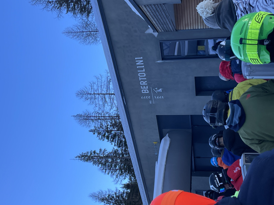 The image, oriented correctly, depicts a winter scene at a ski resort in Courmayeur, Italy, likely near a mountain lodge or a ski lift entrance. A modern-style building with a dark grey facade dominates the right side of the frame. Vertical white lettering on the building clearly displays "BERTOLINI," beneath which are the numbers "1771" and "2277," followed by "mt," suggesting altitudes in meters. A small graphic symbol resembling a person ascending steps is situated above these numbers.
In front of the building, a line of skiers or snowboarders, clad in winter apparel including colorful helmets (notably bright green, black, and orange), wait in what appears to be a queue. Their faces are largely obscured by helmets or turned away. Snow covers the roofline of the building and a curved structure, possibly a barrier for the queue, in the foreground. The background features a clear, bright blue sky and a mix of bare deciduous trees and dark evergreen conifers, indicative of a cold, clear day in the mountains. The overall impression is a bustling yet serene moment at a ski facility.