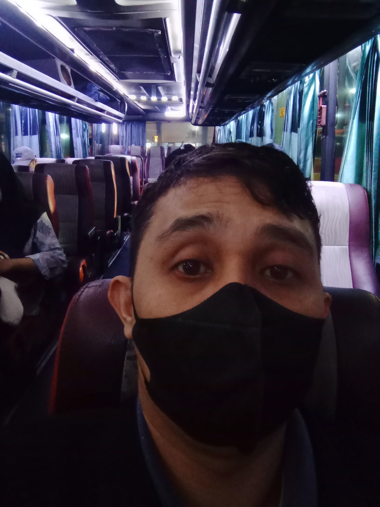 The image shows a close-up selfie of a man wearing a black face mask. He is sitting on a bus, with the interior of the bus visible in the background. The bus has rows of seats with purple and white upholstery, and blue curtains. The man is looking directly at the camera, and his eyes are visible above the mask. The lighting suggests it is either evening or night. The overall scene is consistent with the interior of a passenger bus operating in Surakarta, Indonesia, a city known for its public transportation.