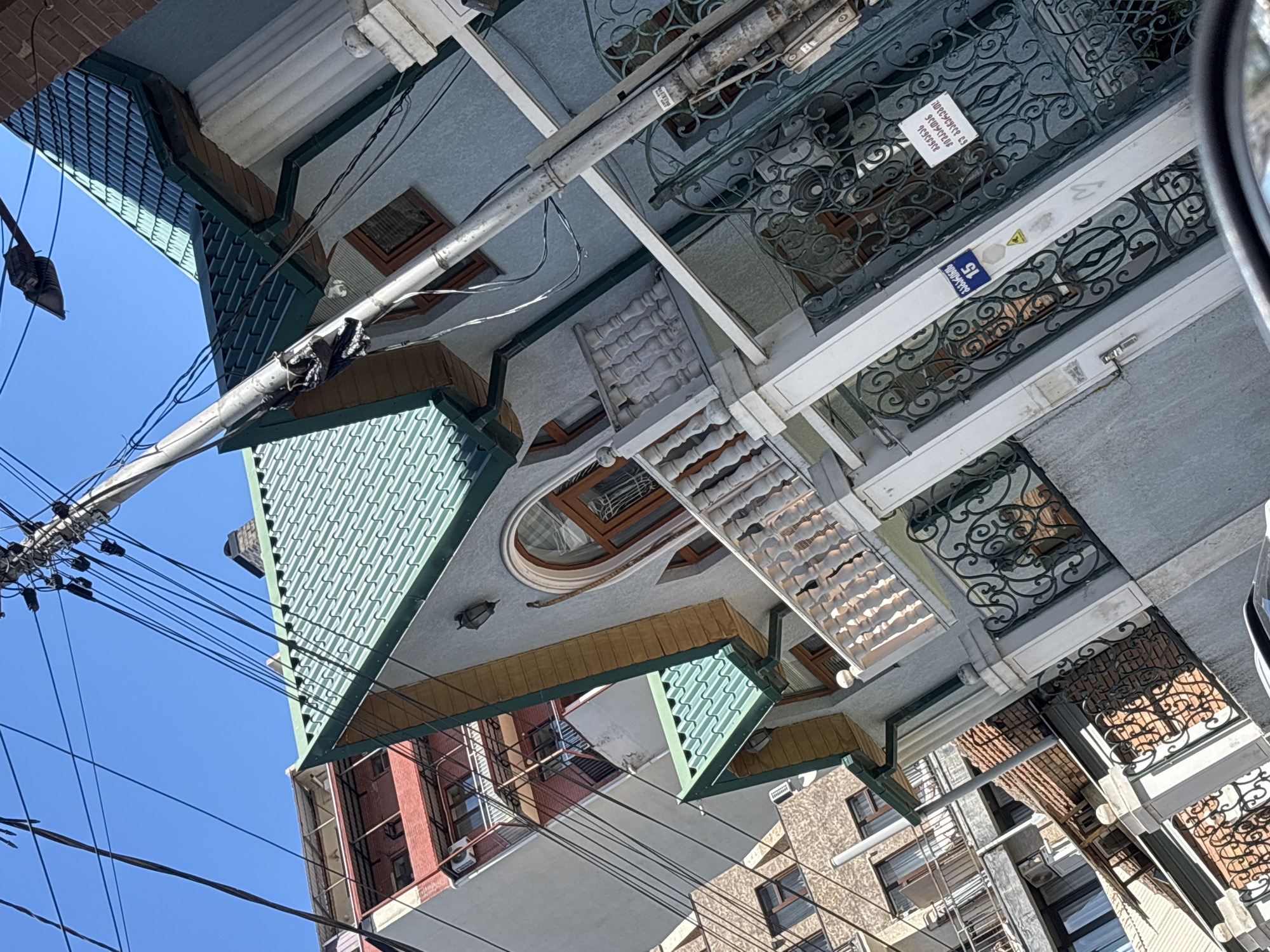 This is a low-angle, close-up photograph of a building on a sunny day, likely in Tbilisi, Georgia, due to the Georgian writing on a sign. The building features a distinctive green, tiled roof with multiple gables. The facade is light blue-grey, and there are ornate wrought-iron gates and a decorative white railing on a balcony. Adjacent buildings, one with a red brick facade and balconies, are visible in the background. A utility pole with a dense network of wires runs diagonally across the frame, obscuring parts of the sky and the buildings. A street number sign with "15" in a blue rectangle is affixed to the gate. Another sign with Georgian writing is also present on the gate. The overall impression is of a well-maintained, residential building within an urban environment.