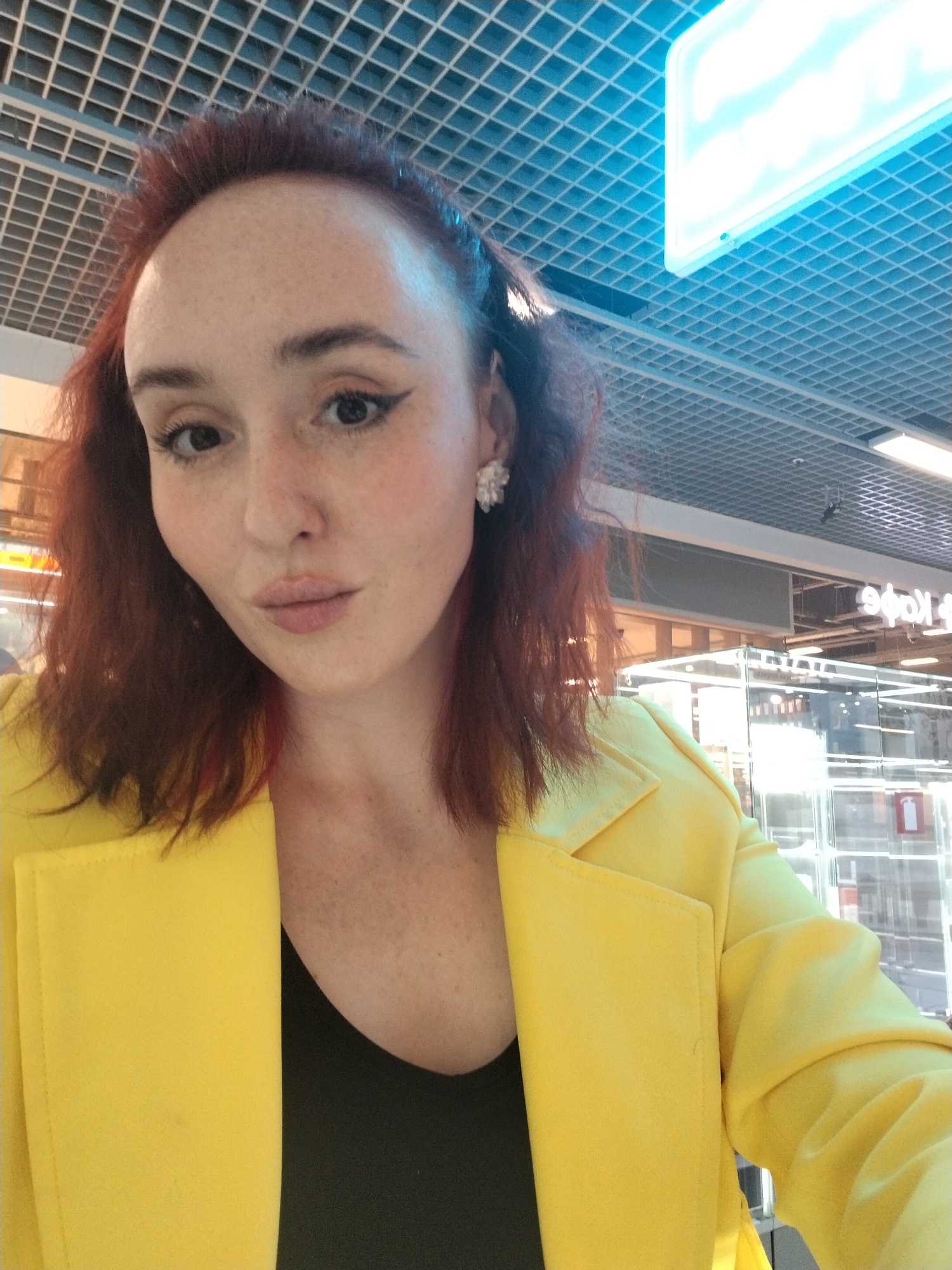 A selfie taken in what appears to be a shopping mall in Izhevsk, Russia. The subject is a fair-skinned woman with red hair and freckles. She is wearing a bright yellow blazer over a black top and floral earrings. She has dark eyeliner and her lips are puckered. The background features a ceiling with a grid pattern and bright lighting fixtures. Storefronts with glass displays and signage in Cyrillic script can be seen in the distance, one reading "Эфа". A fire extinguisher is visible behind her.