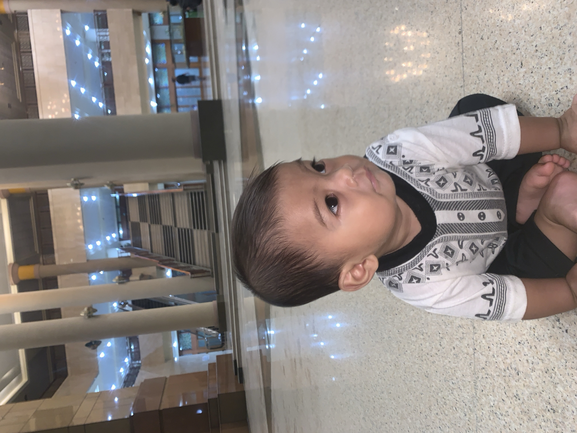A baby is depicted sitting on a light-colored, speckled floor within a spacious indoor environment. The baby, with dark hair and eyes, is wearing a white long-sleeved top featuring black and grey geometric patterns, paired with black shorts or pants. The child is looking upwards and slightly to the right, with a calm and curious expression.
The setting appears to be a large public building, possibly a mall, hotel lobby, or convention center, consistent with a location like Samarinda, Indonesia. The background showcases multiple levels of the building, characterized by polished floors, reflective surfaces, pillars, and railings. Numerous artificial white lights are visible throughout the distant background, casting bright reflections on the floor, suggesting a well-lit indoor space, potentially during the evening or night. The overall atmosphere is clean, modern, and expansive. There is no specific activity beyond the baby observing its surroundings, and no visible text.
