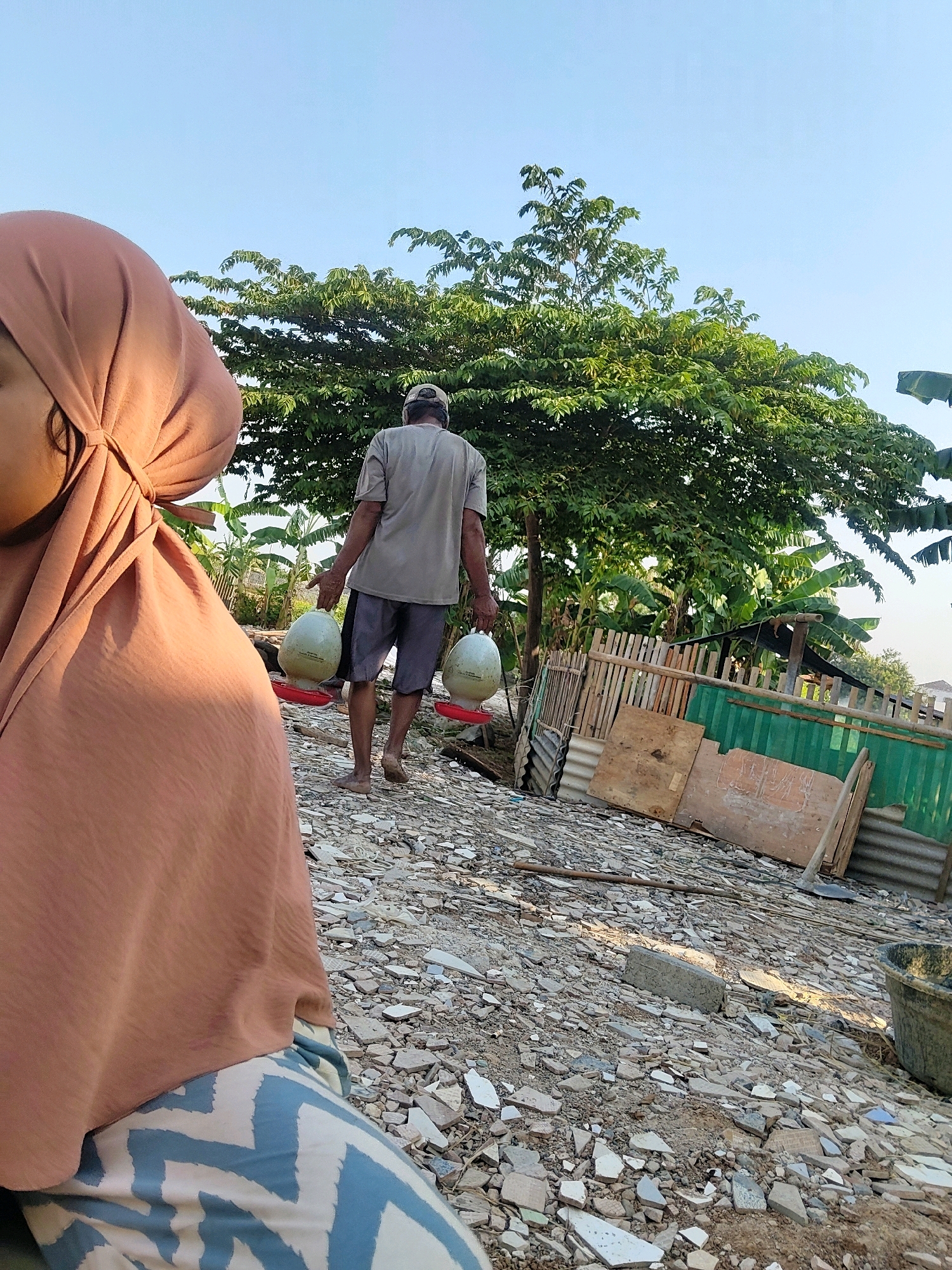 In Mekarmukti, Indonesia, a person wearing a rust-colored hijab and patterned leggings is in the foreground, partially obscuring the view of a man walking away from the camera. The man, wearing a grey t-shirt and dark shorts, carries two large, pale green jugs with red bases in each hand. He is walking on a ground covered with broken tiles and rubble, moving towards a lush green tree. In the background, a wooden fence and some green corrugated metal sheeting are visible, along with banana trees. The sky is clear and blue.