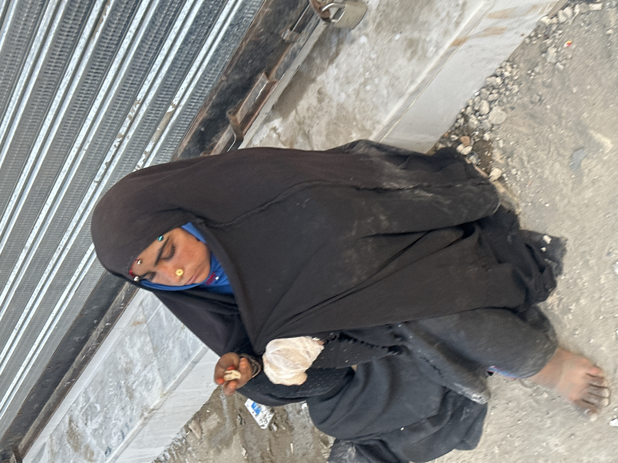 A woman wearing a black burqa sits on a sidewalk in Quetta City Tehsil, Pakistan, beside a closed corrugated metal storefront. She has facial piercings and holds something small in her hands. One foot is bare. The sidewalk is stained and littered with small debris.