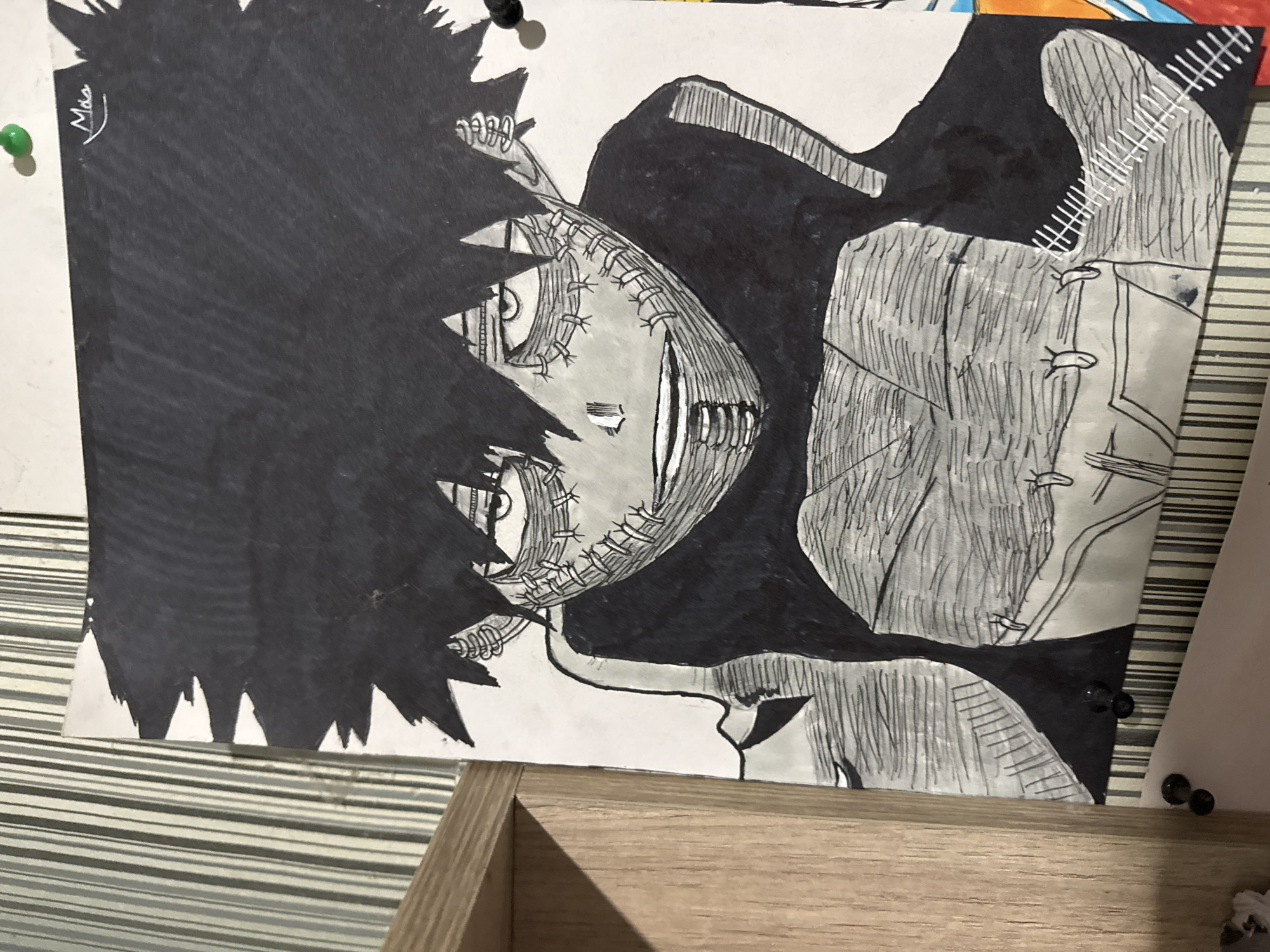 This media file displays a photograph of a monochrome drawing, predominantly featuring an anime or manga-style character. The drawing is rendered in black, white, and various shades of gray, creating a stark visual. The central subject is a character with spiky, dark black hair that covers a large portion of the left side of their head. Their face is pale gray, notable for extensive stitching or staples across the jawline and surrounding the eyes, giving a disfigured or pieced-together appearance. The character has wide, light-colored eyes with dark pupils and displays a wide, somewhat unsettling grin. They appear to be wearing dark clothing, possibly a jacket or cape, with visible texture and additional stitching details on the right shoulder/arm area.
The drawing is on a piece of white paper, which is pinned to a surface. A green pushpin is visible in the upper left, and black pushpins are seen in the upper right and lower right corners, securing the paper. The background surface behind the drawing features a horizontal striped pattern in the upper right and lower left, and a light brown wooden frame is visible in the bottom right corner of the photograph. In the upper left portion of the character's dark hair, a white signature "Mia" is visible. The overall mood conveyed by the character's expression and stitched features is somewhat sinister or unsettling.