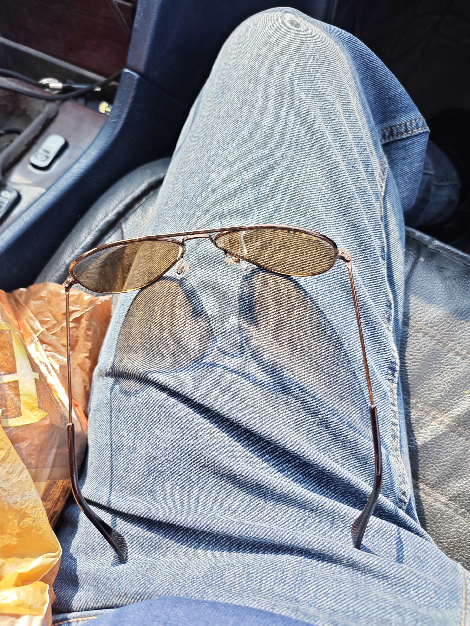 A pair of aviator sunglasses rests on a person's lap. The sunglasses have brown tinted lenses and a thin, copper-colored metal frame. The person is wearing blue denim jeans. The scene appears to be inside a vehicle, with the dark interior of the car visible in the background. To the left of the sunglasses, a portion of a light-colored plastic bag with some visible yellow text is present. The lighting suggests it is daytime, with sunlight casting shadows on the jeans. This image was taken in Vanadzor, Armenia.