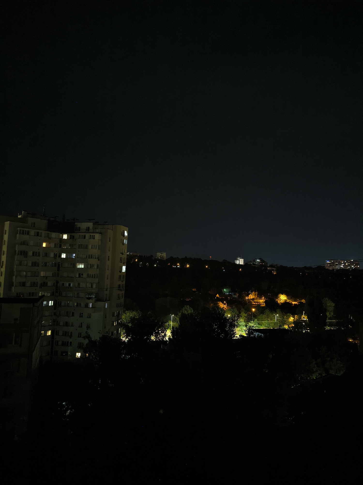 A night-time cityscape, likely in Chișinău, Moldova, is depicted from an elevated vantage point. On the left, a large, multi-story apartment building dominates the foreground, its numerous windows illuminated with warm, yellow light, indicating occupied apartments. The building's facade is light-colored, suggesting it is a concrete structure common in Eastern European cities. In the middle ground and background, a densely wooded area transitions into a hilly terrain. Scattered lights from other buildings and streetlights pierce the darkness, illuminating patches of the landscape and hinting at further urban development beyond the immediate foreground. The sky is a deep, starless black, characteristic of urban night. No people are visible in the image, and the scene is static and devoid of action. The overall impression is one of a residential area at night, with the apartment building serving as the primary focal point against the darker, more natural backdrop.