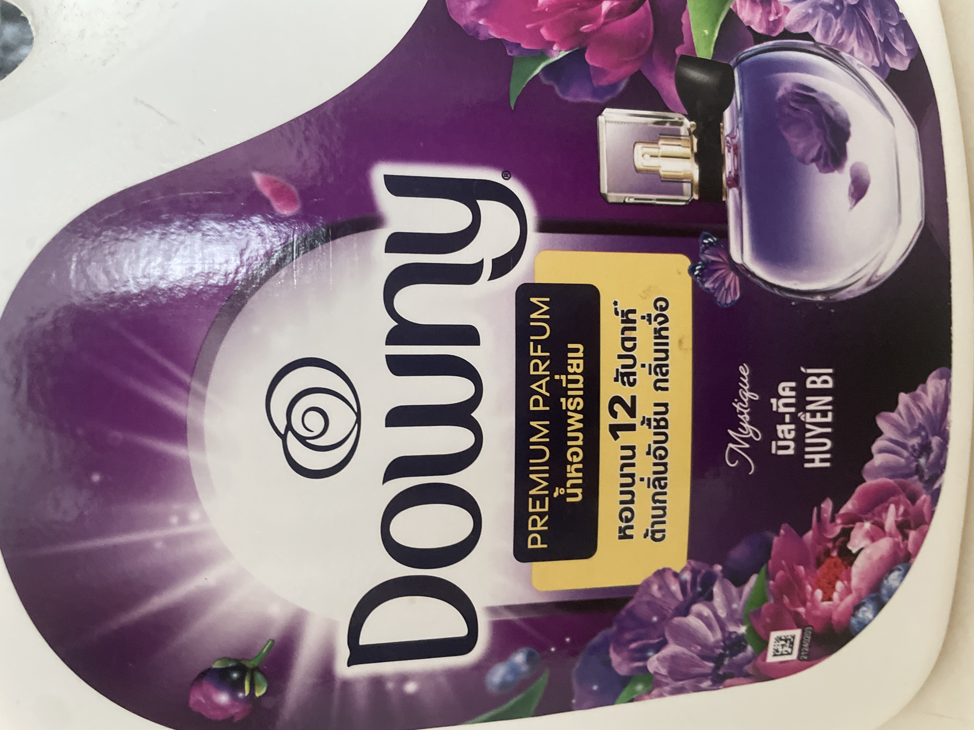 This is a close-up, eye-level, indoor shot of a product label. The label features a prominent "Downy" logo in large, dark blue, sans-serif font. Above the logo is a stylized illustration of a swirled flower. The label's background is a gradient of deep purple, with lighter purple rays emanating from the top center.
To the left of the "Downy" logo, there's an illustration of a small, unopened pink and purple flower bud. The label also includes text in a yellow box. The top line, in bold black text, reads "PREMIUM PARFUM." Below this, in Thai script, it says "น้ำหอมพรีเมียม" which translates to "Premium Perfume."
Beneath the yellow box, in Thai script, there's further text. The first line, in black, appears to be "หอมนาน 12 สัปดาห์" (Long-lasting fragrance for 12 weeks). The second line reads "ต้านกลิ่นอับชื้น กลิ่นเหงื่อ" (Resists musty odors, sweat odors).
On the right side of the label, there's a prominent illustration of a perfume bottle with a purple liquid, a gold cap, and a black bow. The bottle is set against a backdrop of large, vibrant purple and pink flowers, with some scattered smaller flowers and petals. There is also a small purple butterfly illustration on the left side of the perfume bottle.
In the bottom left corner of the label, a QR code is visible, with the number "21240203" below it. Below the perfume bottle illustration, the word "Mystique" is written in a cursive font, followed by Thai script "มิส-ทีค" and then Vietnamese script "HUYỀN BÍ."
The overall impression is a product label for a premium fabric softener or perfume with floral and fruity notes. There are no indications of the city of Pattaya City, Thailand, in this image; it is a product label.