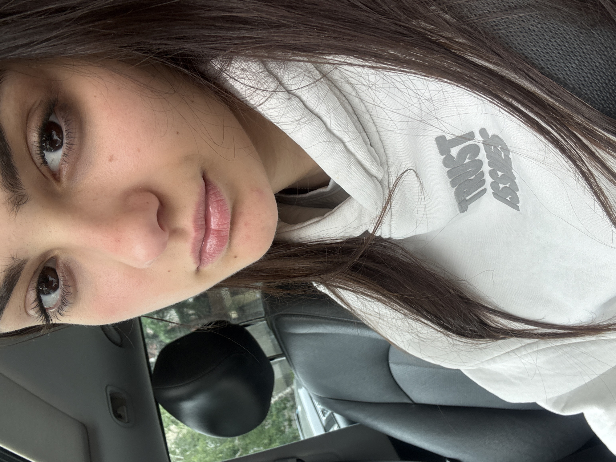 The image shows a woman with dark hair wearing a white hoodie with the text "Trust Issue" on it. She is inside a car, as indicated by the black seat and the car interior visible in the background. The location is unknown and cannot be determined from the image.