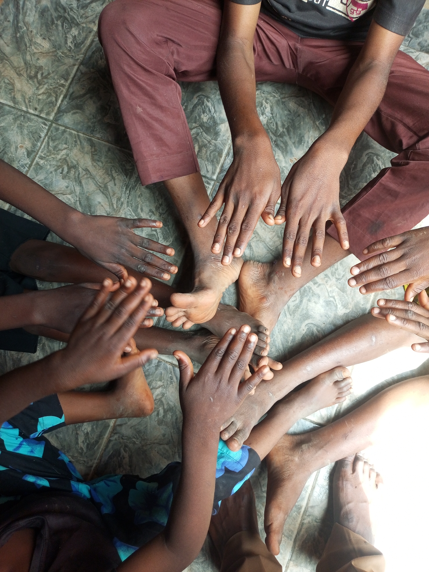 This is a top-down, close-up, forensic photograph of the bare feet and hands of multiple individuals arranged in a circle on a textured, tiled floor. The tiles are gray with a mottled, marble-like pattern. The individuals' skin tones range from deep brown to lighter brown, and their hands are open with fingers spread, some resting on the floor or on other people's feet. One individual is wearing maroon pants, and another is wearing a black t-shirt with a colorful graphic. Another person is wearing a dress with a blue and green floral pattern. The lighting suggests a bright, possibly outdoor or well-lit indoor environment, with a strong light source illuminating the right side of the frame. The scene appears to be a moment of connection or solidarity. The image does not contain specific landmarks or indicators that would allow for precise location identification within Gombe, Nigeria.