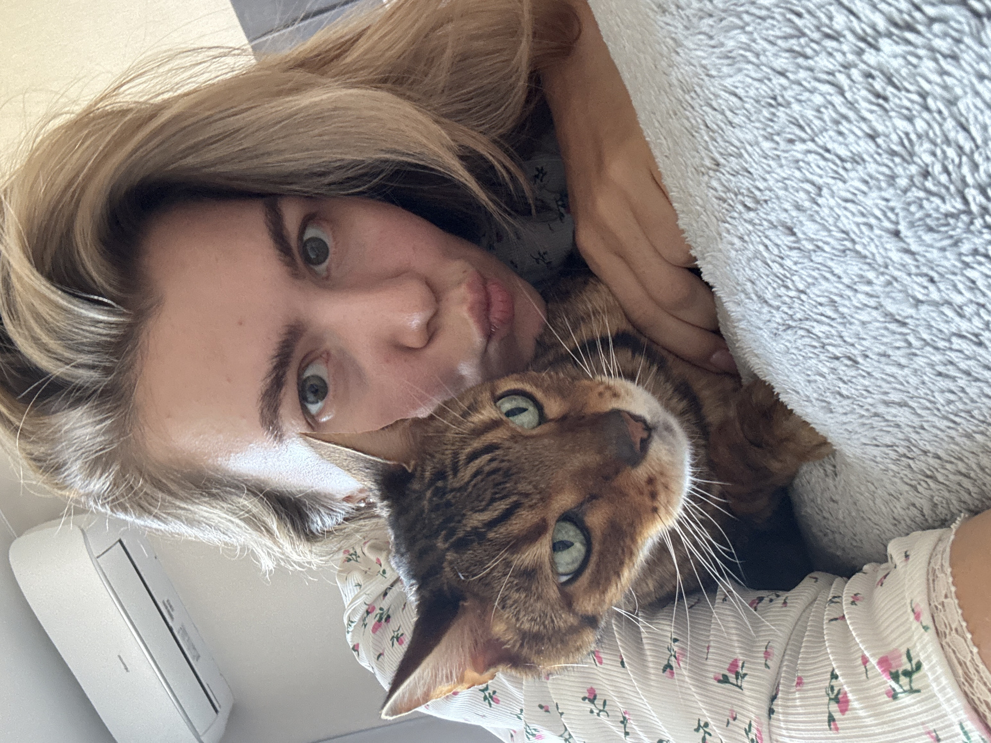 A young woman is captured in a tilted, close-up selfie alongside a Bengal cat in an indoor setting. The woman has light blonde hair and fair skin, with wide eyes and her lips pursed in a playful pout. She is wearing a light-colored top adorned with small pink floral patterns and delicate lace trim. Her arm is partially visible, appearing to hold or cuddle the cat.
The Bengal cat, characterized by its distinctive spotted and marbled brown and black coat, is nestled close to the woman. It looks directly at the camera with striking green eyes and prominent white whiskers. On the right side of the frame, a fluffy, light grey blanket or pillow provides a cozy backdrop. In the background, to the left of the woman's head, a white wall-mounted air conditioning unit is visible. The scene is illuminated by natural light, suggesting it is daytime. The overall mood conveyed is one of affection and a relaxed, playful moment between a pet owner and her cat. No text is visible in the image.