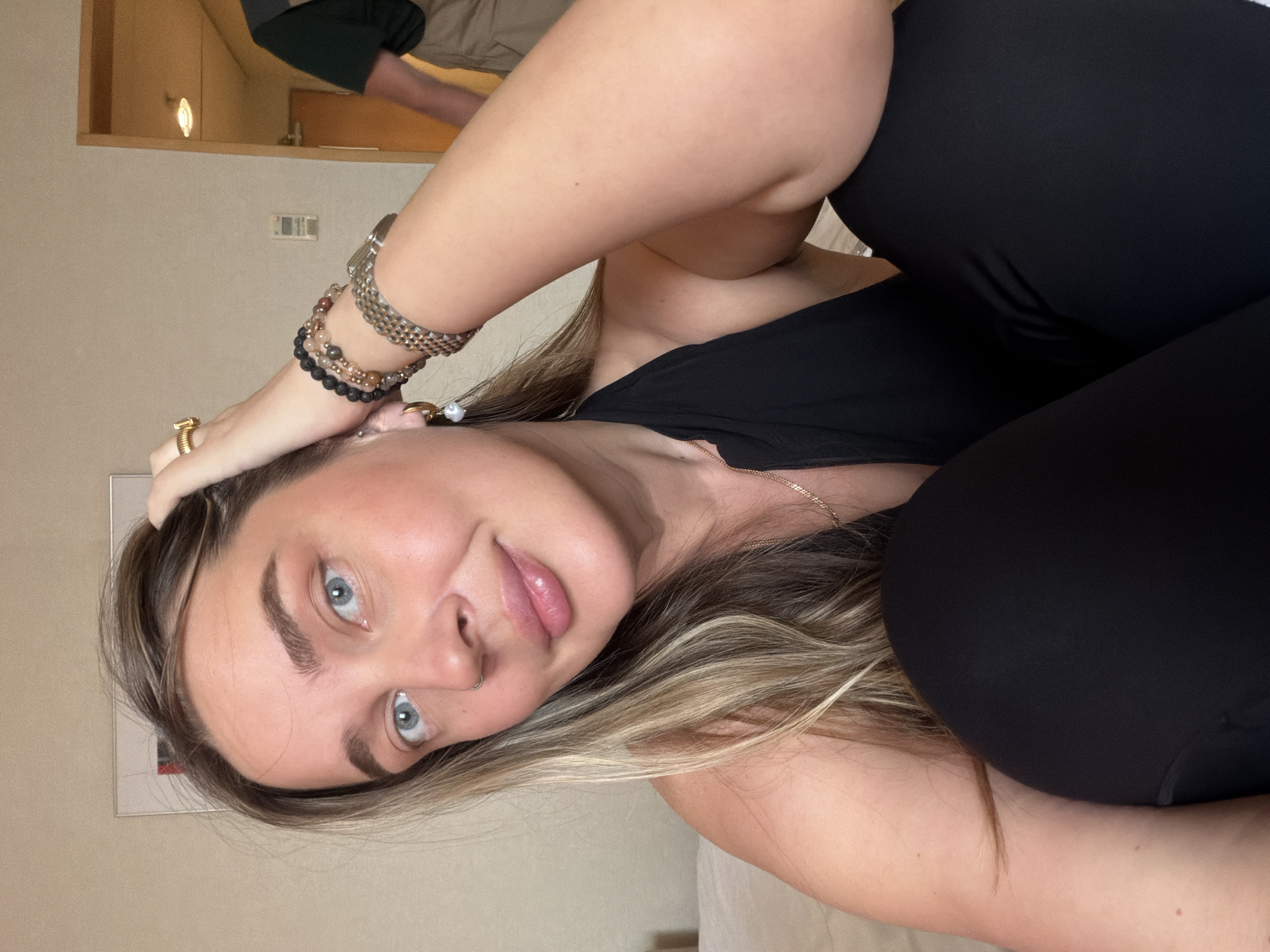 A woman is in a seated or reclined position, angled toward the viewer. She has light skin, blue eyes, and blonde hair. She is wearing a black top and black pants or leggings. She has several bracelets on her left wrist and a ring on her left hand. The background is a room with beige walls. A portion of a man in khaki pants and a dark green or black shirt is visible in the upper left corner. The location is unspecified, and there's no evidence indicating Moscow, Russia.