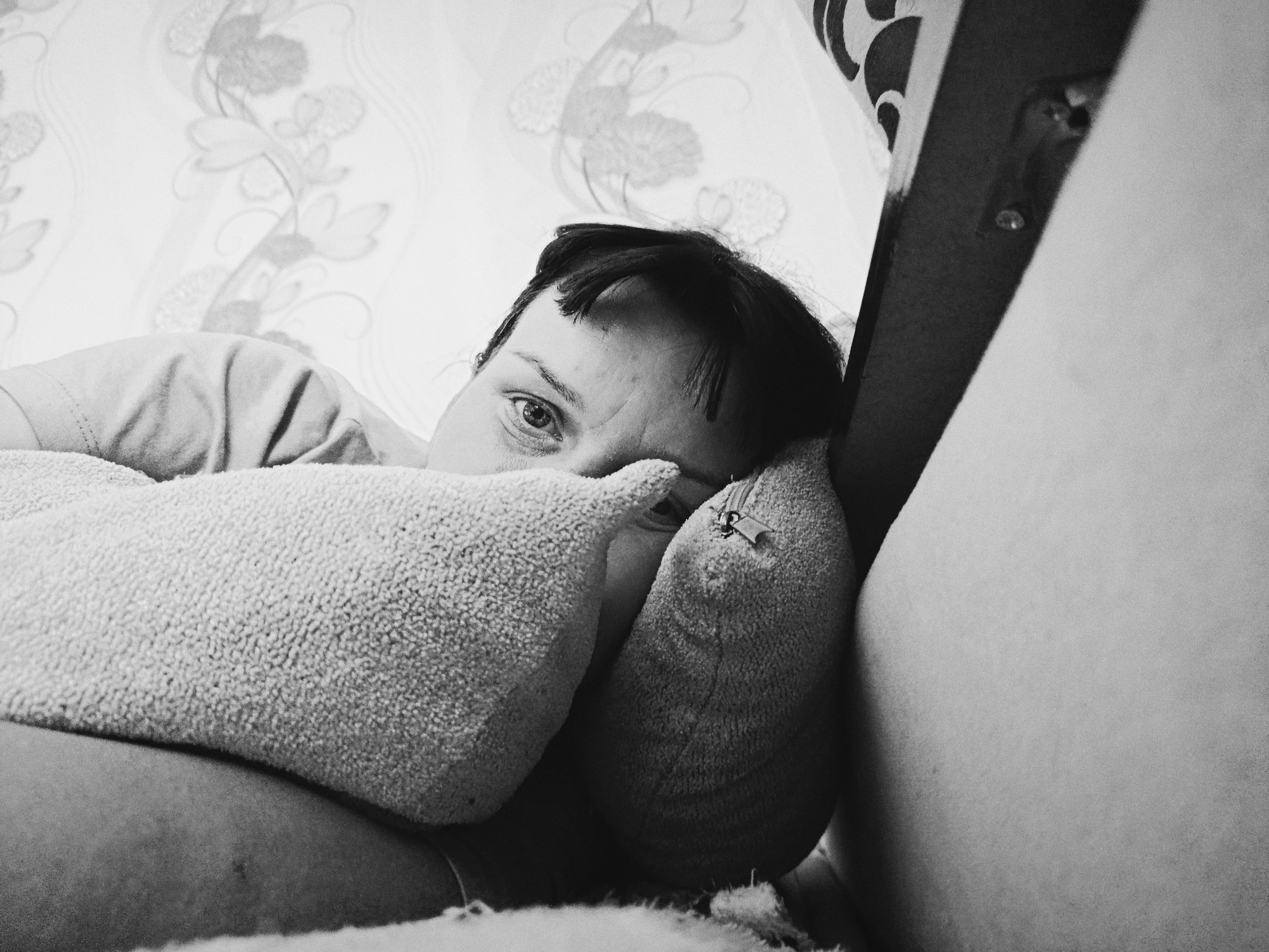 A person is lying on a bed or sofa, partially obscured by two pillows. The person's head is visible, with dark hair and one eye looking directly at the camera. The environment includes a wall with a floral pattern and a corner of a piece of furniture. It is not possible to verify the location as Фоки, Russia, from the image alone.