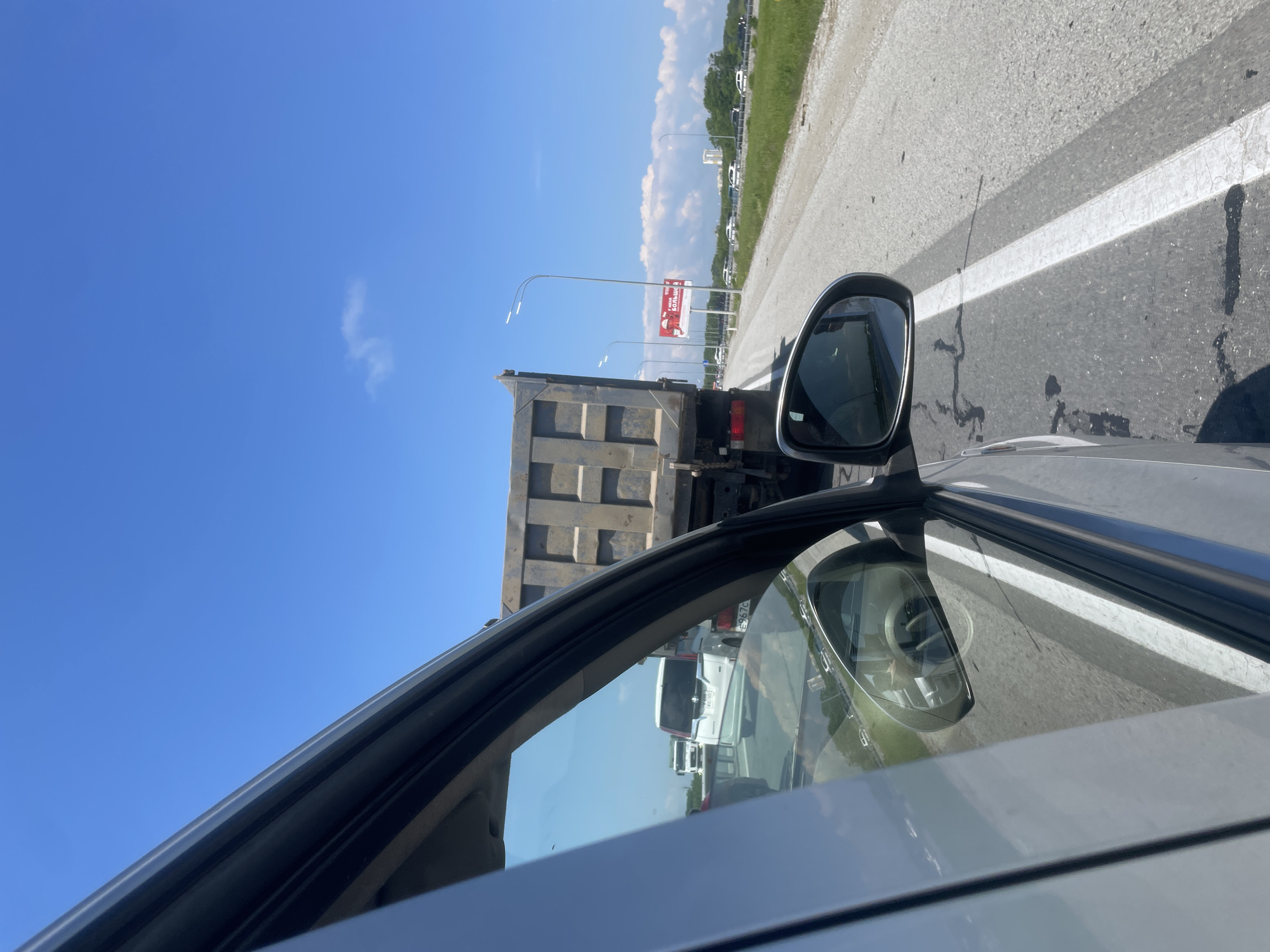 The image shows a view from inside a vehicle, looking out the side window towards a bright blue sky with a few wispy clouds. The foreground is dominated by the interior of the car, with a portion of the door frame and window visible. The side mirror is also in frame, reflecting the road ahead and other vehicles.
The main subject is a large, empty dump truck directly in front of the camera vehicle, in what appears to be a traffic jam on a highway. Beyond the dump truck, other vehicles can be seen, including a white minivan and a white truck further down the road. The highway has a clear white lane marking and is bordered by a grassy verge. In the distance, there are trees and some buildings, suggesting an urban or semi-urban environment. A billboard with Russian text and a red graphic is visible on the right side of the road. The overall impression is a sunny day on a busy road, possibly with a traffic delay. The location is identified as Solovej-Kljuch, Russia, suggesting this is a view of a highway or major road within or near that locality.