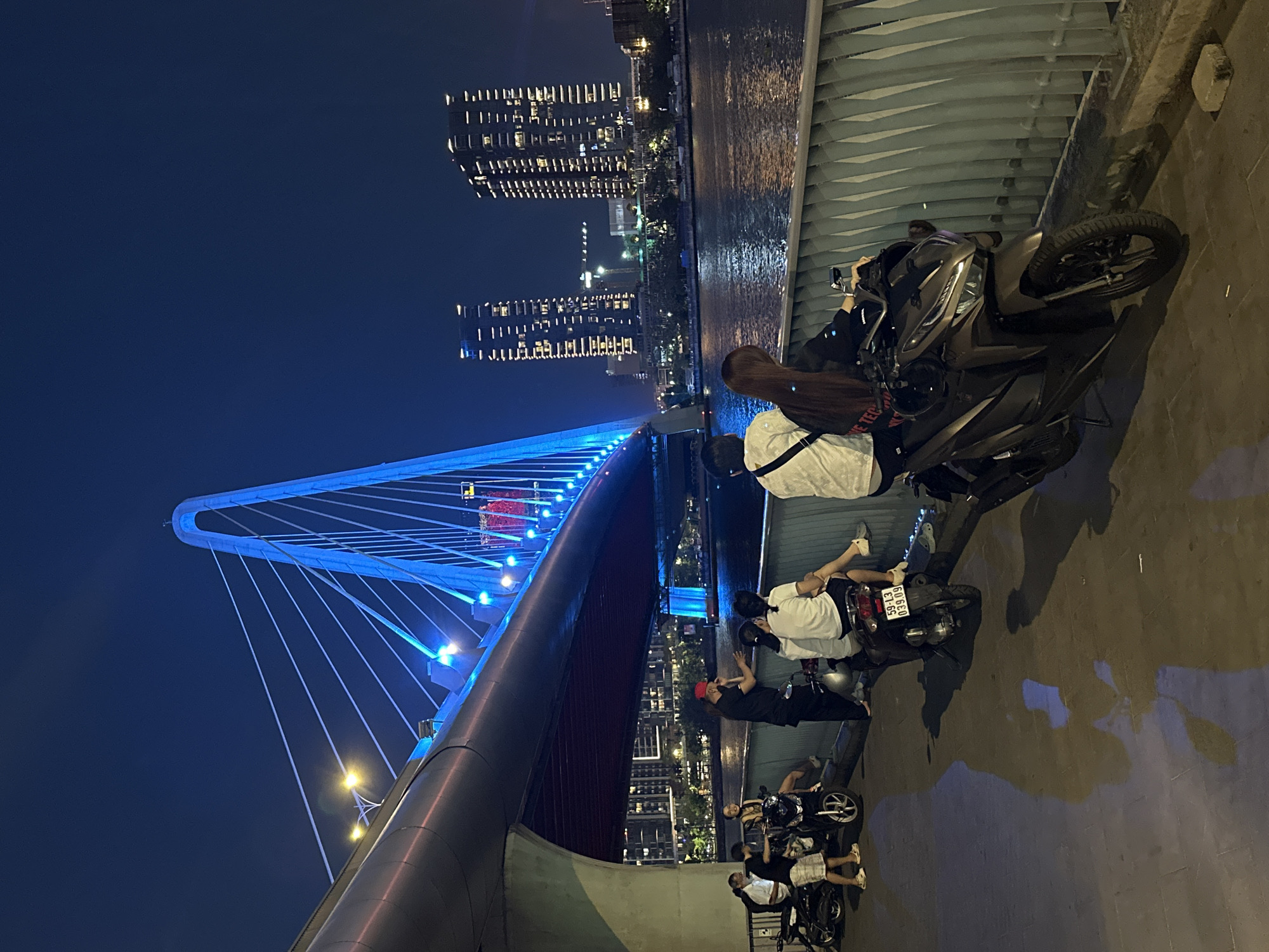 At night, a bridge in Thủ Đức, Vietnam, is illuminated in blue, with tall buildings in the background. Several people are gathered on the bridge near motorcycles, some sitting on the bikes. They appear to be socializing and enjoying the view. The environment includes the bridge structure, the city skyline with illuminated buildings, and the dark sky.