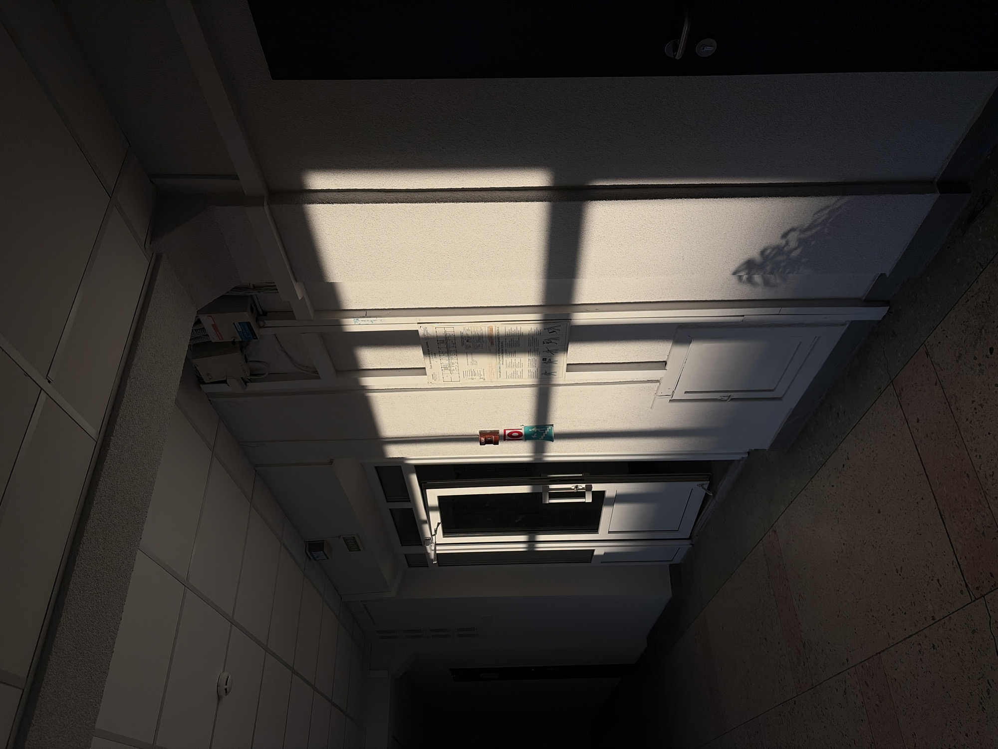 This media file captures an interior architectural space, likely a hallway or corridor, characterized by stark contrasts of light and shadow. The image's perspective is angled, looking upwards or across a wall.
The central feature is a light-colored, textured surface, possibly a ceiling or high wall, bathed in a strong beam of natural light. This light casts prominent, sharply defined geometric shadows, suggestive of a window frame, across the surface. Alongside these, an intricate shadow resembling tree branches or foliage is distinctly visible, implying an external natural element.
Positioned below the main illuminated area is a white panel structure. Several paper documents with illegible text and diagrams are affixed to this panel. Nearby, three small square signs are visible: two red, and one blue-green featuring a white running figure, typically indicating an emergency exit. Integrated into this panel structure is an open, rectangular hatch or window, revealing a dark interior beyond and equipped with a handle.
To the left, a section of a light-colored, paneled wall with vertical divisions and a textured column is visible. The right side of the frame shows a darker, mottled surface, likely a tiled floor or a lower wall section, receding into shadow. The top and bottom edges of the image are filled with dark, undefined architectural surfaces.
The sharp, angled shadows indicate direct sunlight, placing the time of day as daytime, possibly morning or late afternoon. The scene is devoid of people and conveys a functional, institutional, or commercial building environment. The only decipherable text is the iconic symbol on the emergency exit sign.