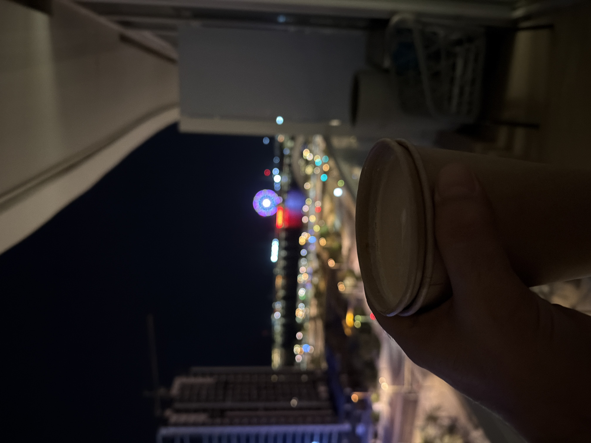 A first-person perspective shot captures a hand holding a disposable coffee cup filled with a light-colored beverage. The cup is a neutral tan color and is held in the foreground. In the background, the night cityscape of Unknown, Unknown unfolds, illuminated by numerous colorful lights. Tall buildings, a construction crane, and a Ferris wheel are visible, along with a body of water. The scene appears to be taken from a balcony, with the edge of the structure and part of an outdoor chair visible on the right side.