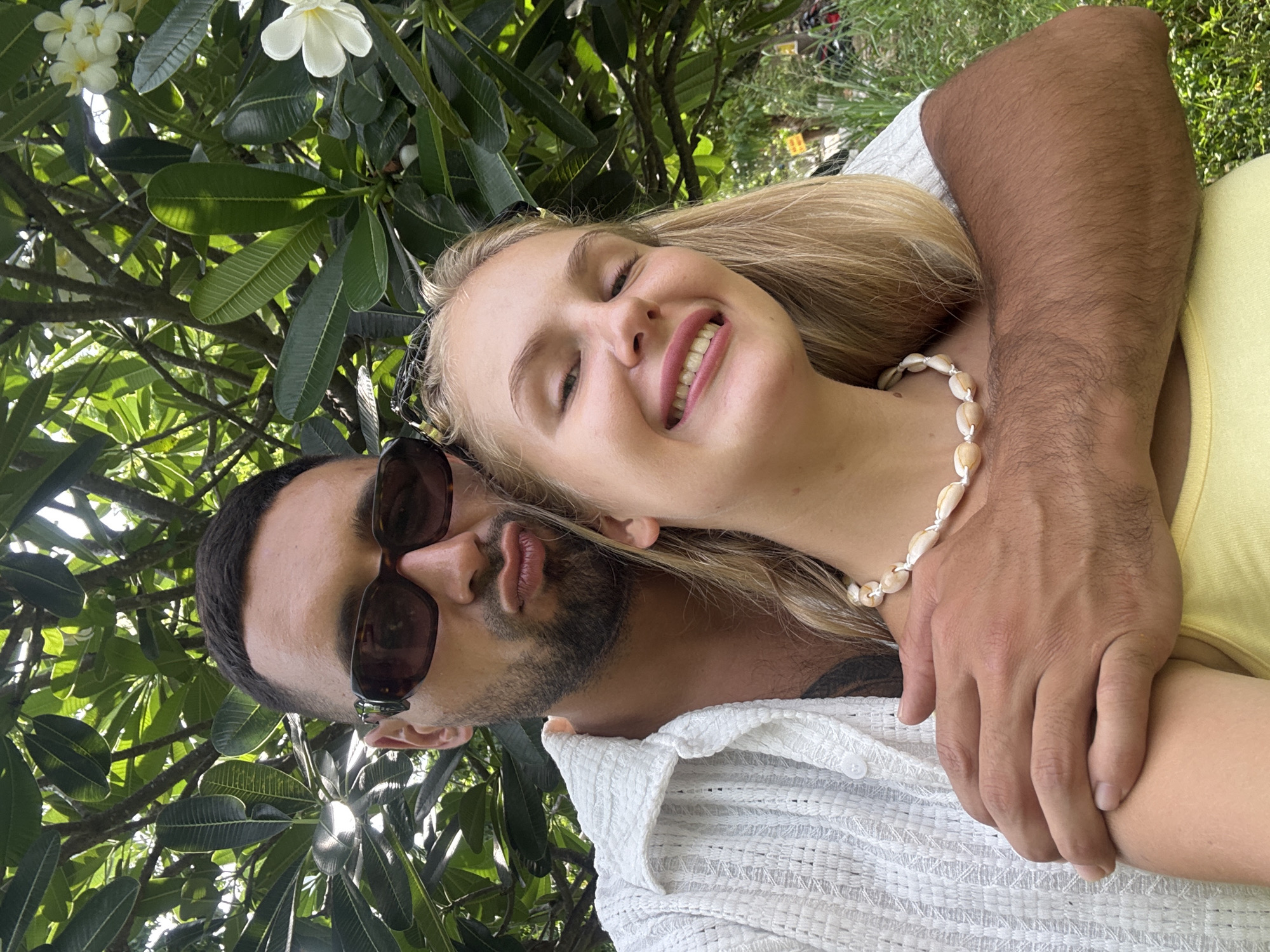 A man and woman pose for a selfie in a lush, green outdoor environment, likely a park or garden in Nam Nha Trang Ward, Vietnam. The man, positioned on the left, is wearing dark sunglasses and a white textured button-down shirt. He has short, dark hair and a beard, and his lips are puckered. His arm is around the woman's shoulder. The woman, on the right, has blonde hair and is wearing a yellow top and a shell necklace. She is smiling broadly at the camera, her eyes are partially closed. Behind them, large green leaves and white flowers are visible, suggesting a tropical or subtropical setting. The lighting indicates it is daytime.