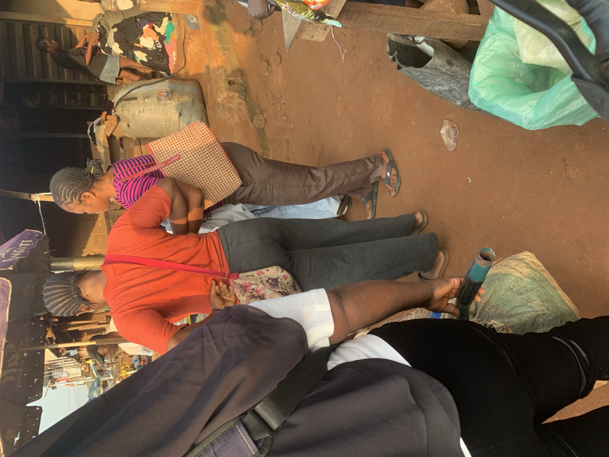 This is a medium shot, eye-level, natural light, outdoor photograph taken in the city of Shagamu, Nigeria. The scene depicts a bustling market setting. In the foreground, a person wearing black pants and a dark blue shirt is partially visible from the side, with a black strap bag across their chest. To their right, a woman in an orange t-shirt and grey pants stands facing away from the camera, with a floral-patterned bag slung across her shoulder. Further right, another woman in a pink and black striped shirt and brown pants is also facing away, carrying a woven bag. In the background, several large sacks are visible, along with other market stalls and people. The ground is a reddish-brown dirt. The overall impression is one of everyday commerce in an African urban environment.