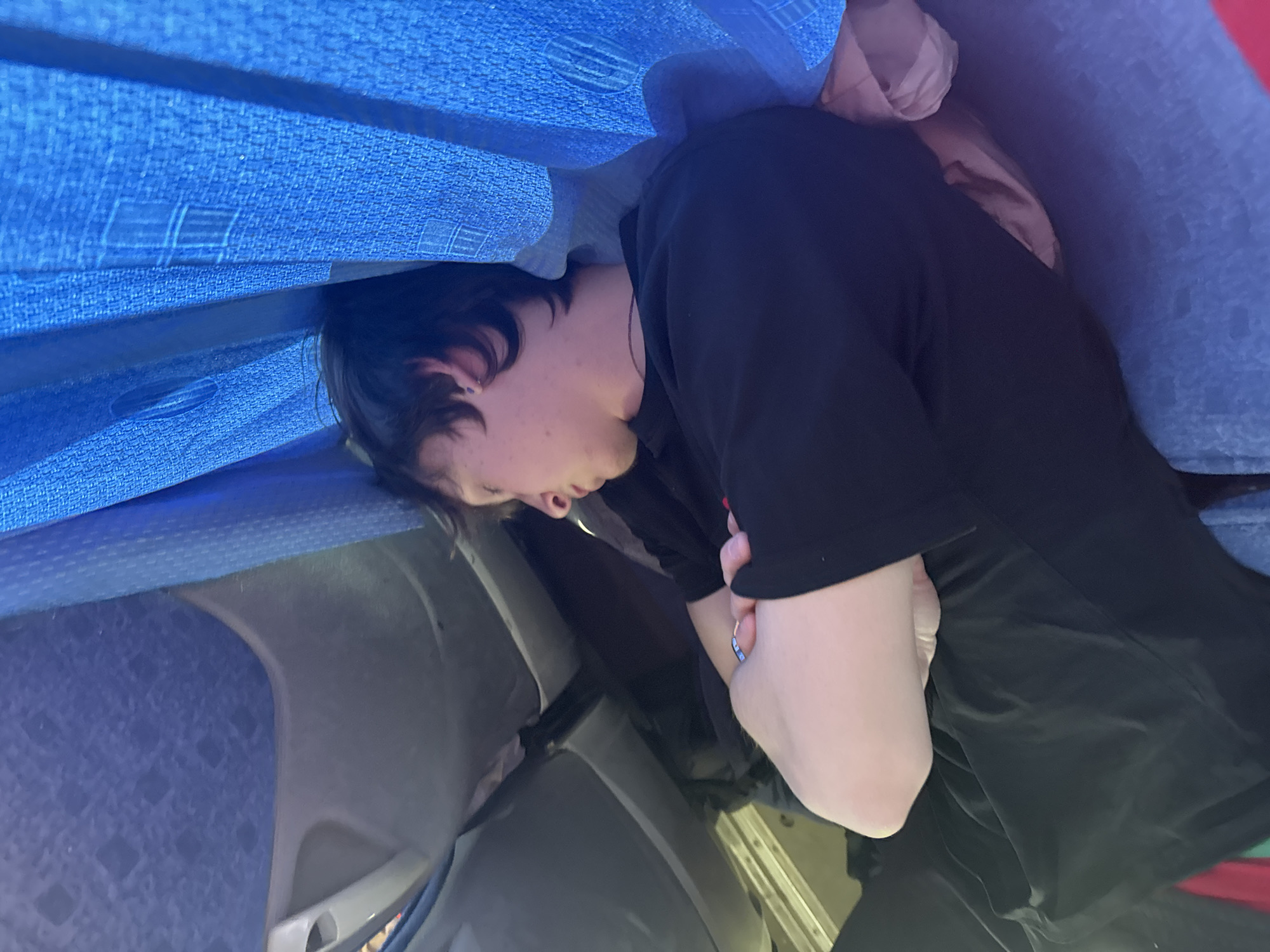 A person is sleeping on a bus. The person has dark hair and is wearing a black shirt. The bus has blue seats and curtains. The location is Челва, Russia.