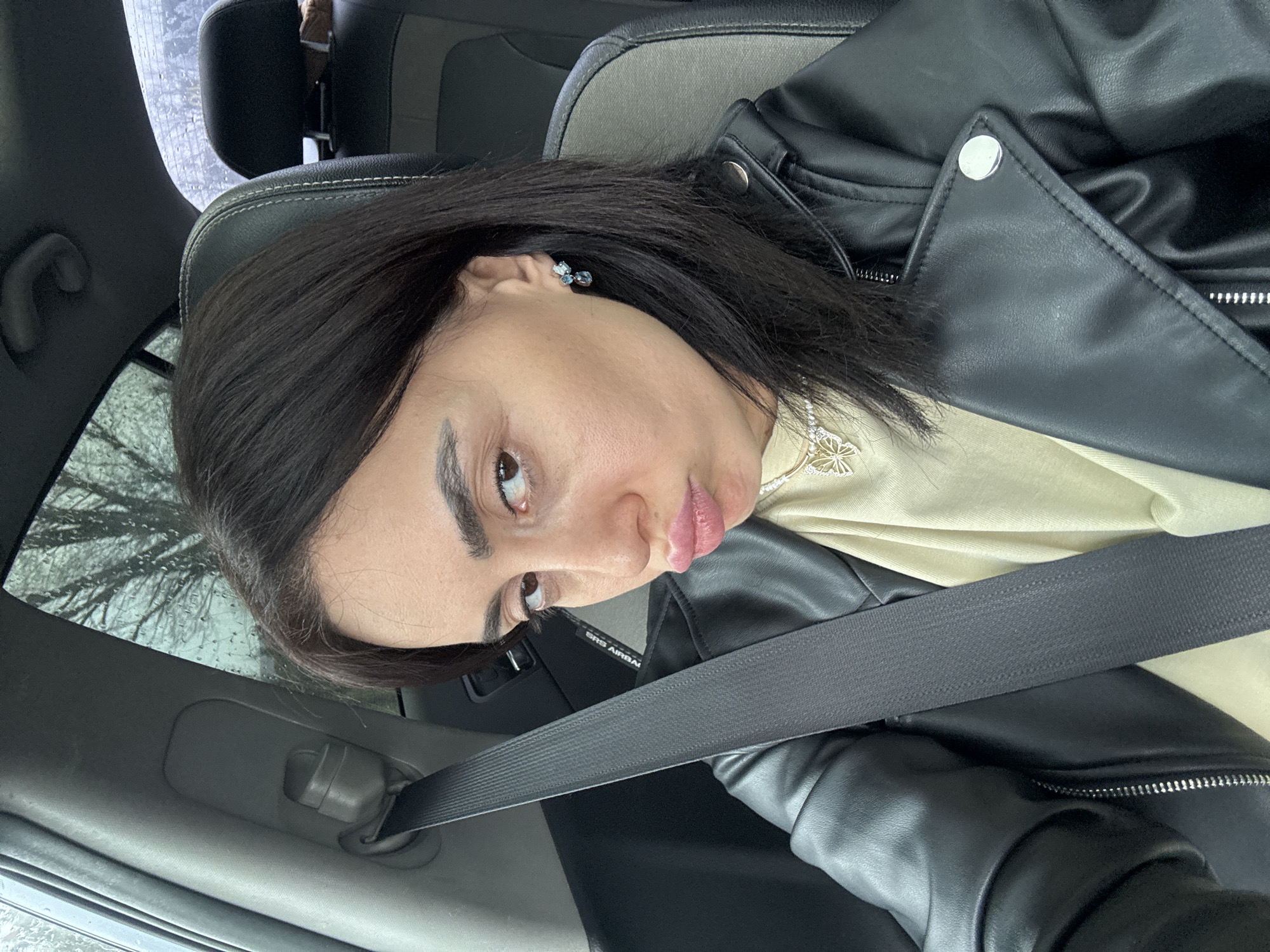 A female individual is depicted in the front passenger seat of a vehicle, secured by a black three-point seatbelt. She has dark hair, dark eyebrows, and brown eyes, looking towards the camera with pursed lips. She is wearing a black leather jacket over a light-colored top, a necklace featuring a gold-toned butterfly pendant, and earrings with blue and clear stones. The vehicle's interior features black seats with gray inserts and stitching. Through the side and rear windows, visible water droplets indicate rain, and bare tree branches are discernible in the background. An "AIRBAG" label is visible on the passenger side panel. The scene is situated within a vehicle in the city of Tbilisi, Georgia.