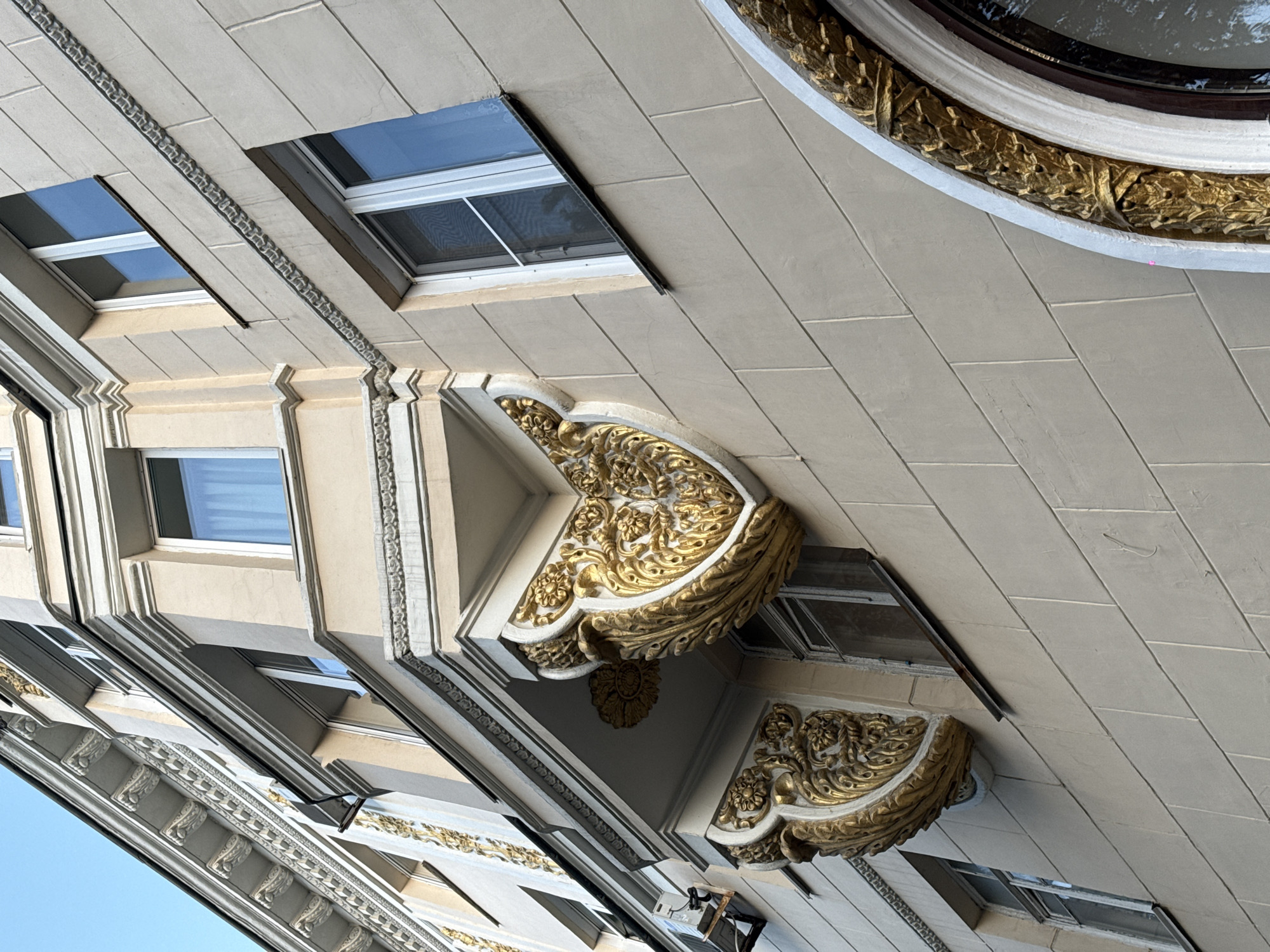 This is a low-angle, close-up shot of the exterior of a building in Chelyabinsk, Russia. The building features ornate golden architectural details, including decorative elements resembling floral motifs and leaves, applied to a light-colored, possibly stuccoed, facade. There are several windows visible, some with white frames and blue-tinted glass, and others with screens. Two prominent decorative elements are suspended from balconies or ledges, each appearing as a gold-colored, heart-shaped or shield-shaped ornament with intricate scrollwork and floral designs. A portion of a large, arched window with a similar golden leaf-patterned frame is visible in the lower right of the image. The sky is a clear blue. The architectural style suggests it could be from the late 19th or early 20th century, possibly Art Nouveau or a similar decorative style.