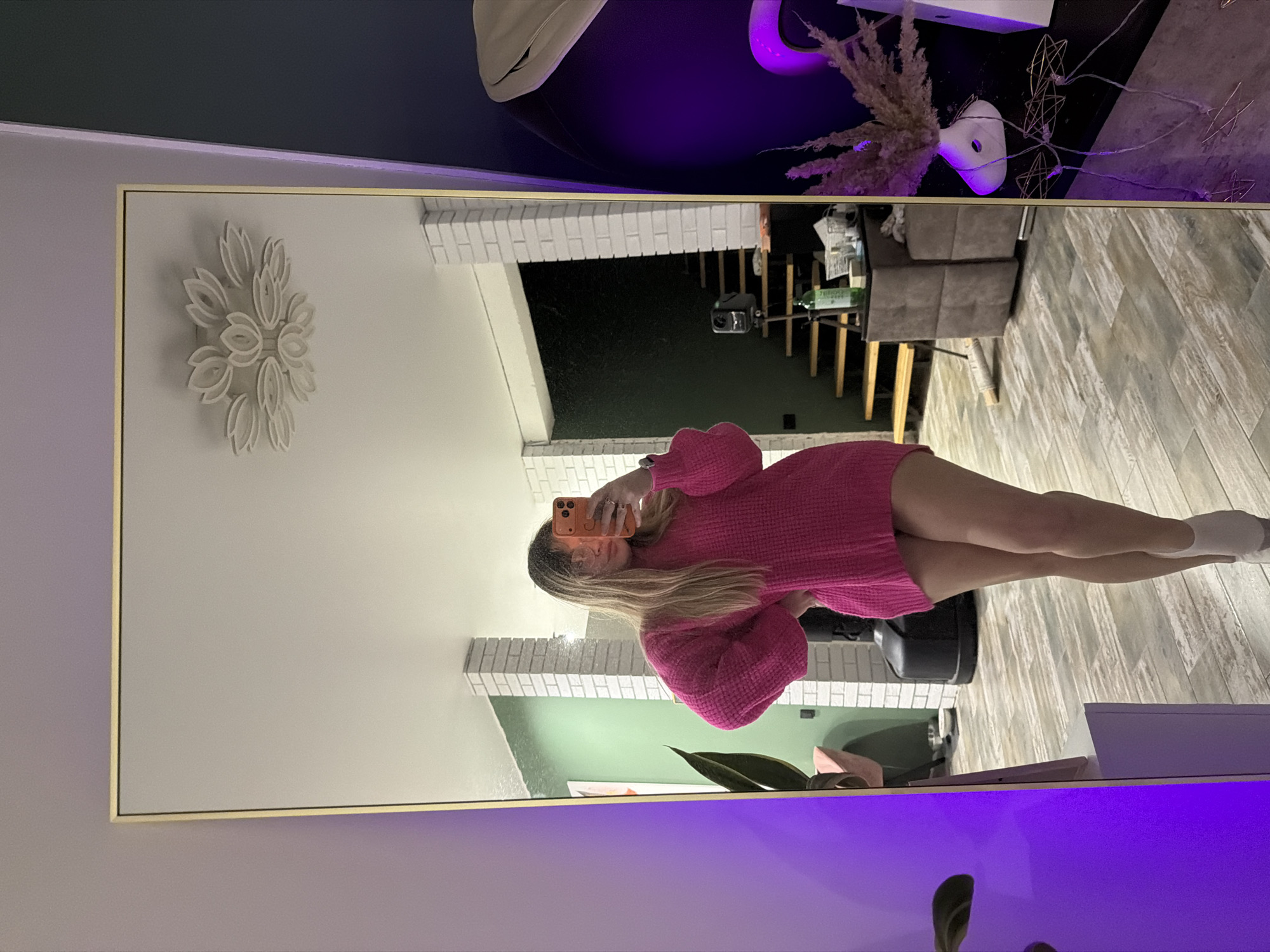 A reflection in a large mirror dominates the frame. In the reflection, a woman stands facing the mirror, holding a smartphone up to take a selfie. She is wearing a pink, oversized sweater and white socks. The reflection also reveals the room's decor, including a white brick wall, a green wall, and a white flower-shaped decoration on the wall.
Behind the woman in the reflection, a short flight of stairs can be seen leading up to another level of the house. There's a table with items on it, and part of a sofa is visible. The floor appears to be made of light-colored wood planks.
The actual room where the mirror is located has purple lighting. Objects partially visible in the room include a vase with dried flowers and the top of a plant.
The location is Большая Шильна, Russia, implied from metadata.