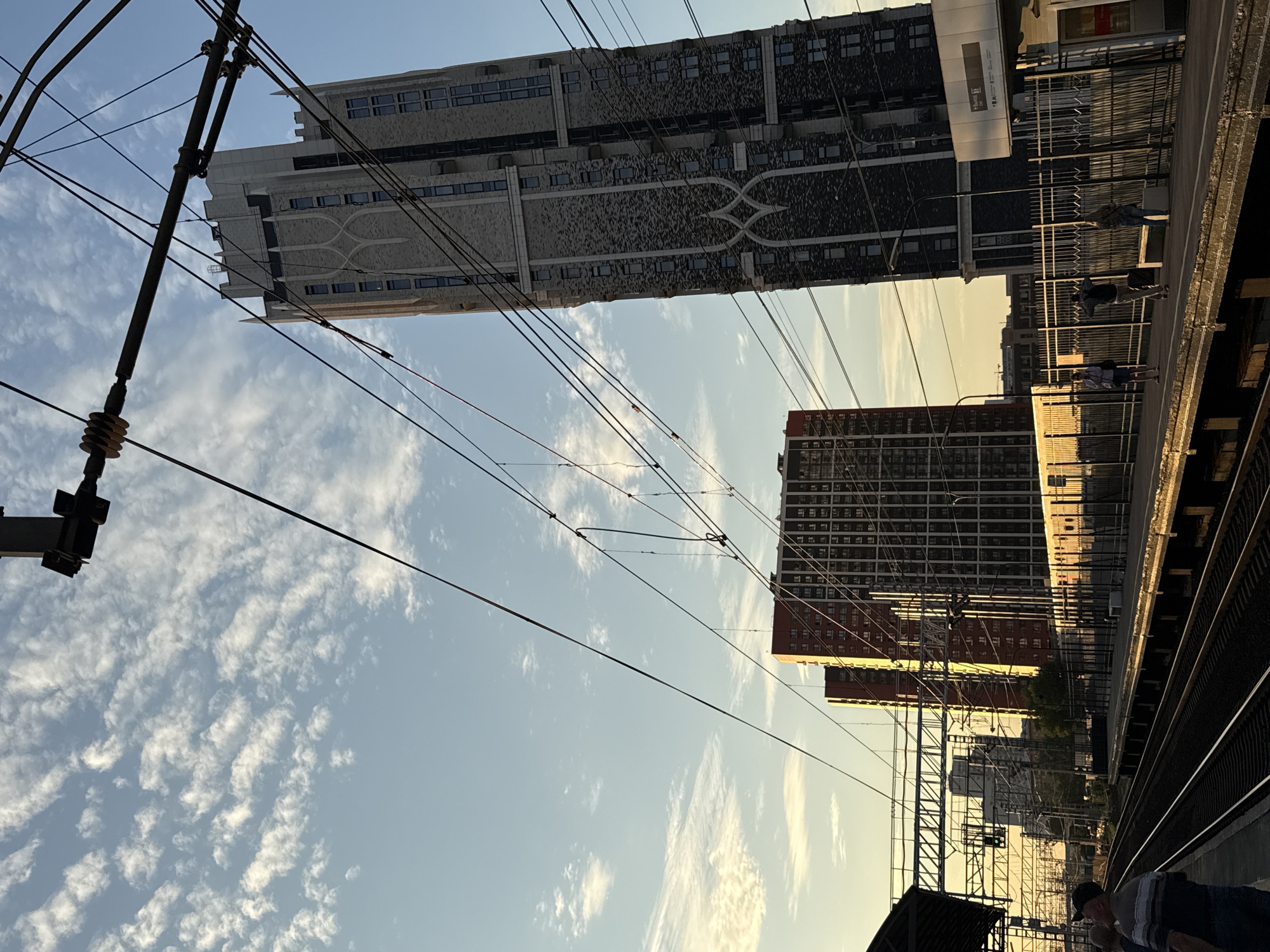 A low-angle shot captures a view of a train platform and tracks with several high-rise buildings in the background. The sky is a pale blue with wispy white clouds and is illuminated by the warm glow of what appears to be sunset or sunrise. Overhead, a complex network of train power lines and supporting structures crisscrosses the frame.
On the train platform, several individuals are visible. To the left, two men are standing on the platform, looking towards the tracks. One man wears a striped shirt and jeans. Further down the platform, three people are visible, standing and appearing to wait. One person is wearing a blue shirt and dark shorts, another a plaid shirt and khaki pants, and the third a grey long-sleeved shirt and jeans. A bench is also present on the platform.
In the background, three distinct buildings stand out. The closest one to the right is a tall, slender building with a dark facade featuring a repeating pattern and numerous windows. To its left, a slightly shorter, red-brick building with many windows is visible. To the left of the red-brick building, a lighter colored high-rise building is also present.
The train tracks stretch into the distance, with the platform running alongside them. The overall scene suggests an urban environment with active transportation infrastructure.