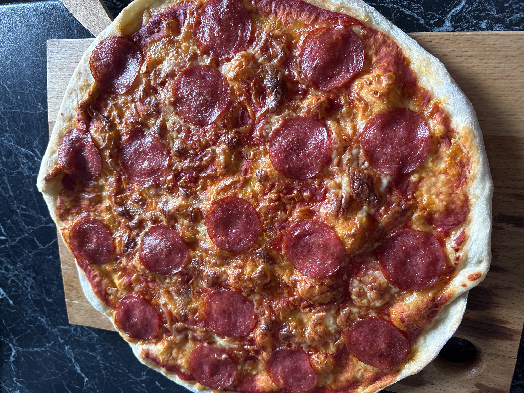 This close-up, overhead shot captures a freshly baked pepperoni pizza resting on a light wooden cutting board. The round pizza occupies the majority of the frame, showcasing its golden-brown crust and generous toppings. Numerous slices of bright red pepperoni are evenly distributed across the melted cheese and tomato sauce base. The cheese is bubbly and golden, indicating it's hot and freshly cooked, with some areas slightly crisped and browned. The edges of the pizza crust are light golden, with a slightly puffed appearance.
The wooden board extends partially into the frame, positioned on a dark countertop that features a subtle white, marble-like pattern. The lighting is bright and even, suggesting it is daytime indoors, likely in a kitchen or dining area in Hatne, Ukraine. There is no activity depicted, but the pizza appears ready to be served. No text is visible in the image. The overall impression is one of a warm, inviting, and appetizing meal.