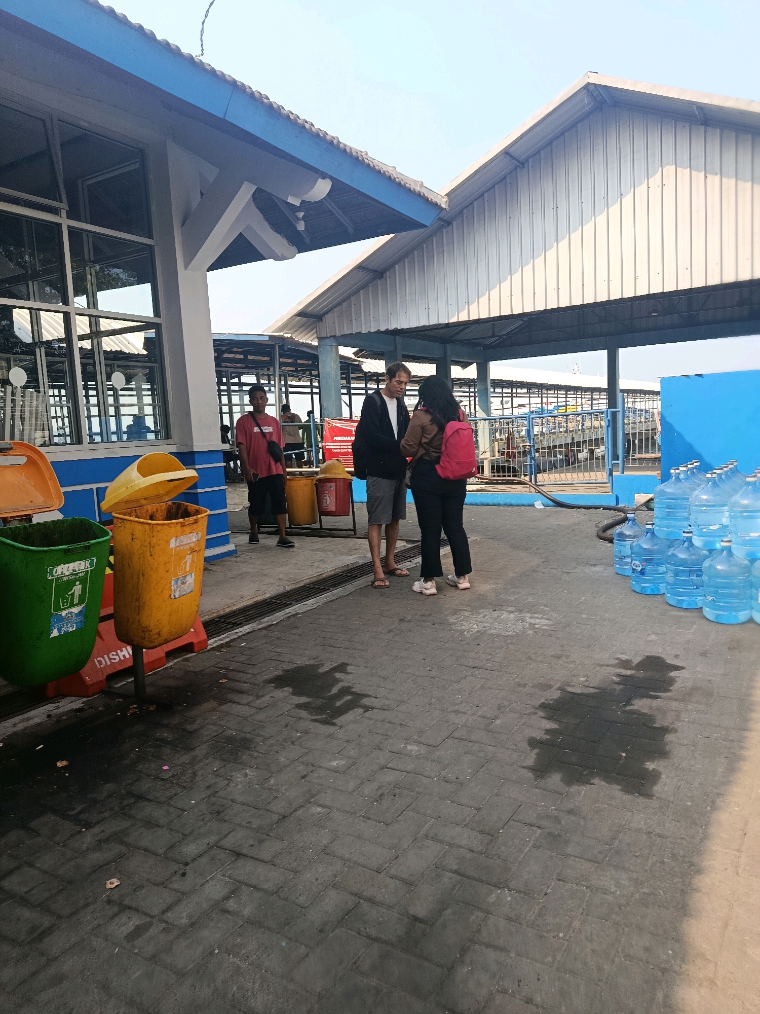 The image depicts a scene at a ferry terminal in Jepara, Indonesia. In the foreground, three large trash bins are visible: a green one on the left, an open orange one in the middle, and a yellow one on the right. A man in a red shirt and black shorts, wearing a black crossbody bag, stands to the right of the trash bins. Further to the right, a couple stands facing each other, engaged in conversation. The man is wearing a black jacket and grey shorts, while the woman is wearing a brown top and black pants, with a red backpack. Behind them, a row of large blue water jugs are stacked. The background shows the ferry terminal structure with a blue and white roof. In the distance, a ferry is docked. The ground is paved with interlocking bricks, and there are some water puddles on the ground. The lighting suggests it is daytime, possibly late afternoon, given the shadows.