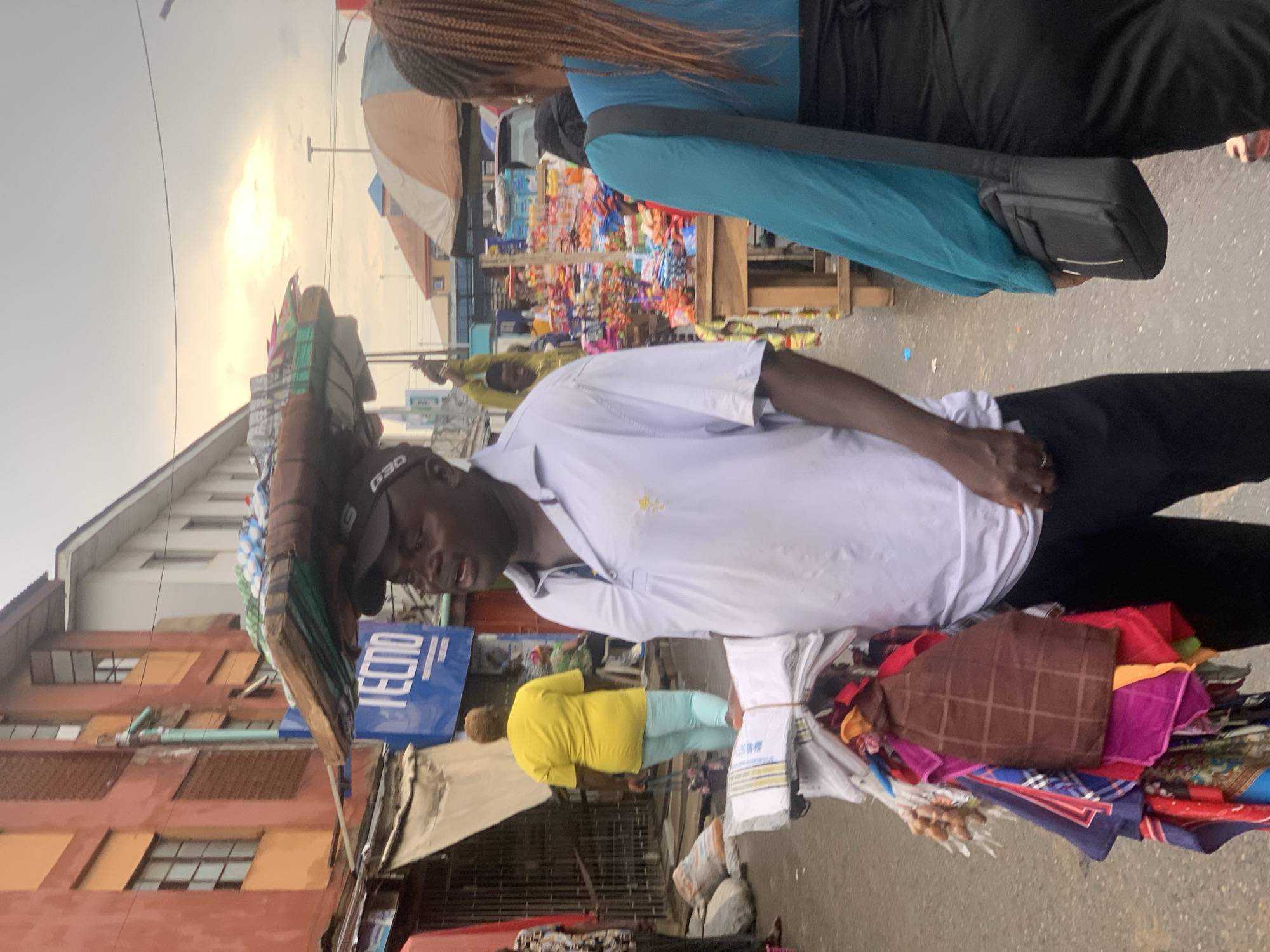 The image captures a street scene in Ikorodu, Nigeria. A man is carrying a large stack of goods on his head. He wears a white collared shirt, black pants, and a baseball cap. He is looking down. Near him stands a person with braided hair in a blue shirt and dark pants, with a black bag over their shoulder. The background features a crowded market area with shops and vendors. There are buildings along the side of the street and market vendors can be seen in the background.