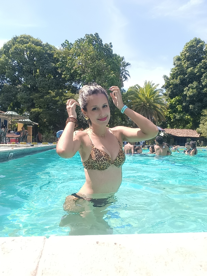 An outdoor scene depicts a light-skinned adult female standing waist-deep in a turquoise-blue swimming pool under a clear blue sky. She possesses light-colored, wet hair and looks directly at the camera with a slight smile. She wears a leopard-print push-up bikini top and dark-colored bikini bottoms. Her arms are raised, with her hands positioned near her temples. Accessories include a gold-colored necklace, gold-colored earrings, multiple brown beaded bracelets on her right wrist, and a light blue wristband on her left wrist.
The foreground features the white tiled coping of the swimming pool. In the mid-ground, several other individuals are partially submerged in the pool behind the primary subject. To the left background, a shaded area with at least two green patio umbrellas and tables is visible, with other people seated or standing. Lush, dense green trees, including palm trees, fill the upper background, indicating a tropical or subtropical environment. The scene is brightly illuminated by direct sunlight. The depicted location is an outdoor recreational pool facility, specified as being in La Victoria, Venezuela.