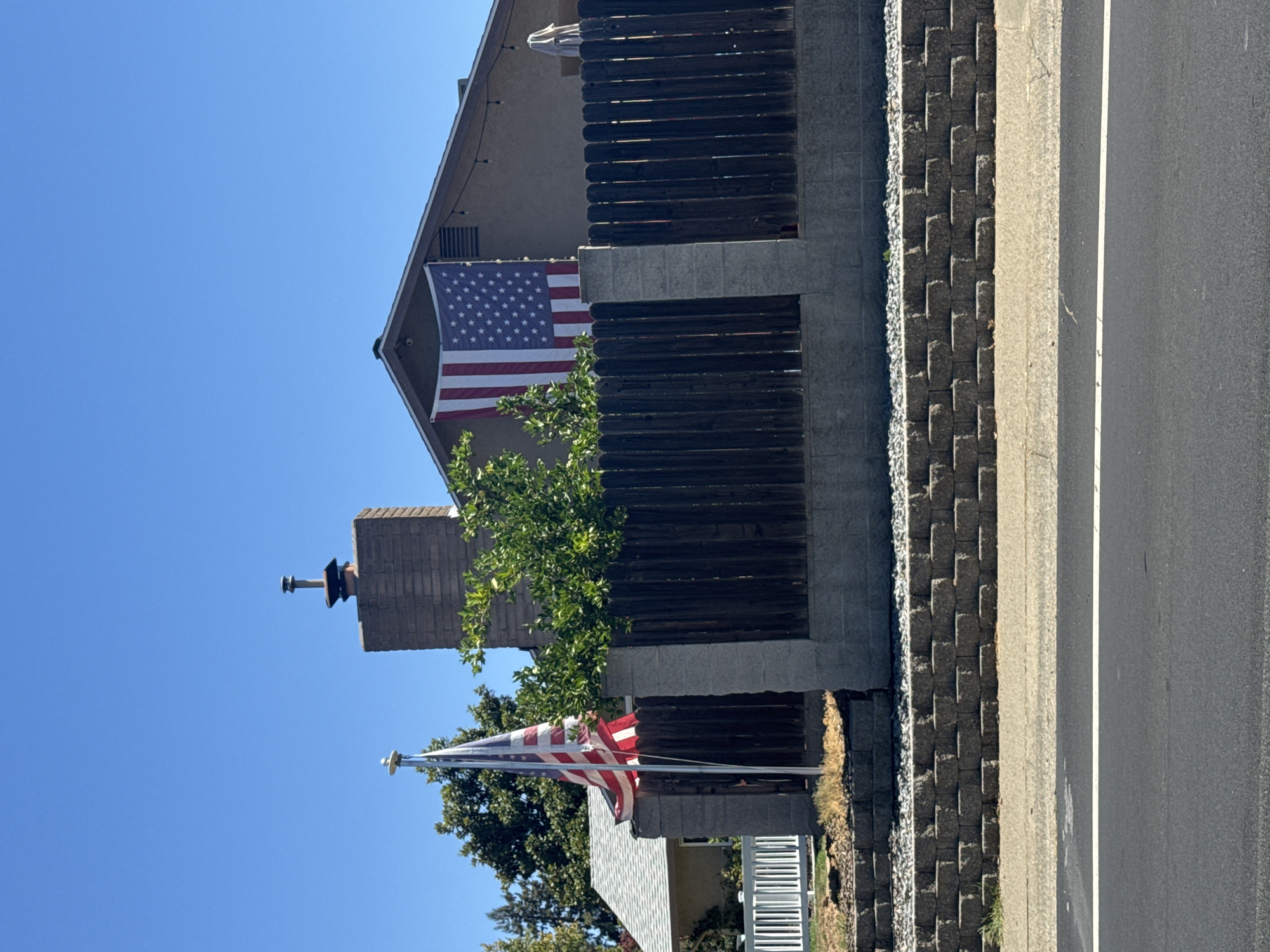 The image shows an American flag flying from a flagpole and another American flag draped over the eave of a house. The house is located in Roseville, United States of America, and has a brick chimney. A wooden fence with a concrete base runs across the foreground. The sky is clear and blue. There is a road in front of the fence.
