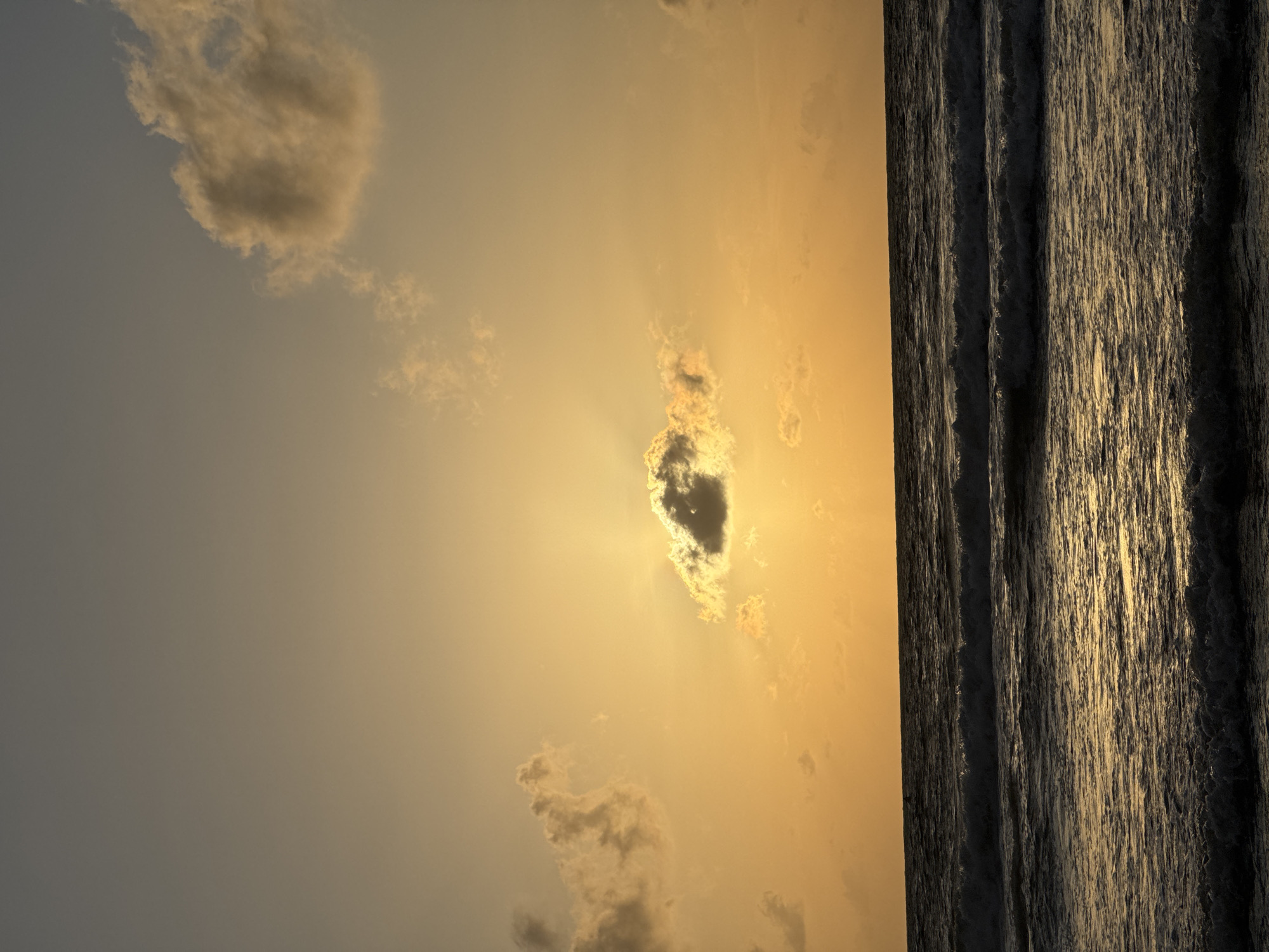 The image depicts a sunset over the ocean in Patong, Thailand. The sky is filled with hues of orange and yellow, with a few scattered clouds. One prominent cloud is positioned in the center of the frame, partially obscuring the setting sun, which casts rays of light through its edges. The ocean is calm, with gentle waves rolling towards the shore. The water reflects the warm colors of the sky, creating a shimmering effect. There are no people or man-made structures visible in the image. The overall scene is serene and picturesque, capturing the natural beauty of a sunset on a Thai beach.
