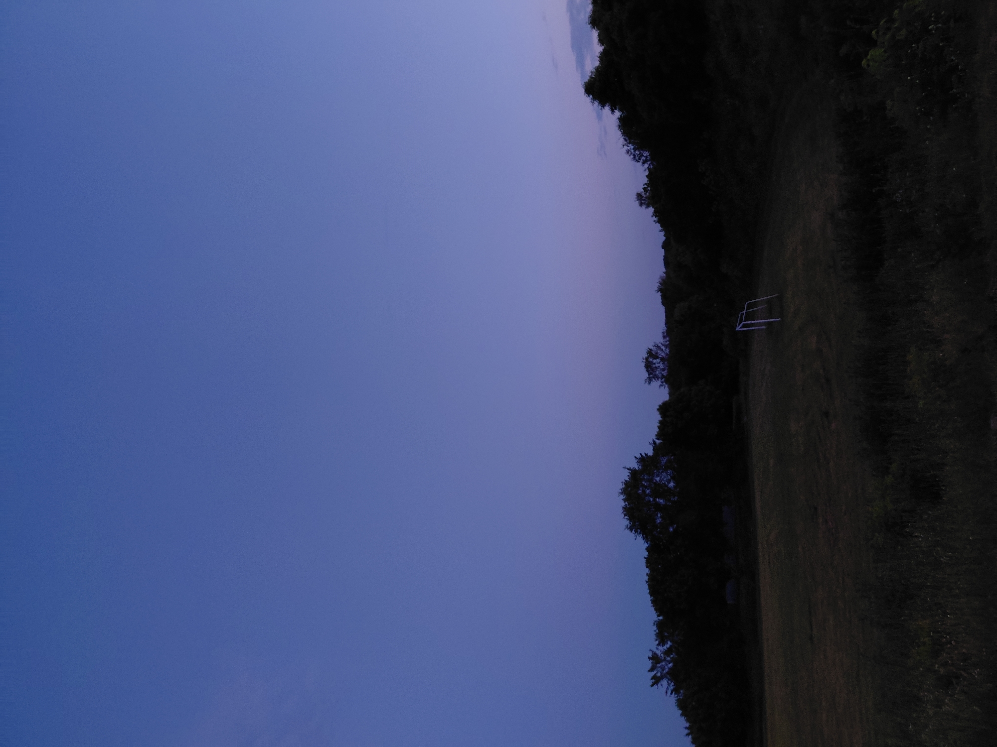 The image depicts a soccer goal on a grassy field at dusk. The sky is a deep twilight blue, transitioning to a lighter shade near the horizon. Dark silhouettes of trees form a backdrop behind the field. The soccer goal is white and appears to be made of metal, positioned on the left side of the field. The grass is somewhat overgrown and uneven. The overall scene is peaceful and quiet, characteristic of a rural or park setting. The location is stated as Ustyvytsia, Ukraine.