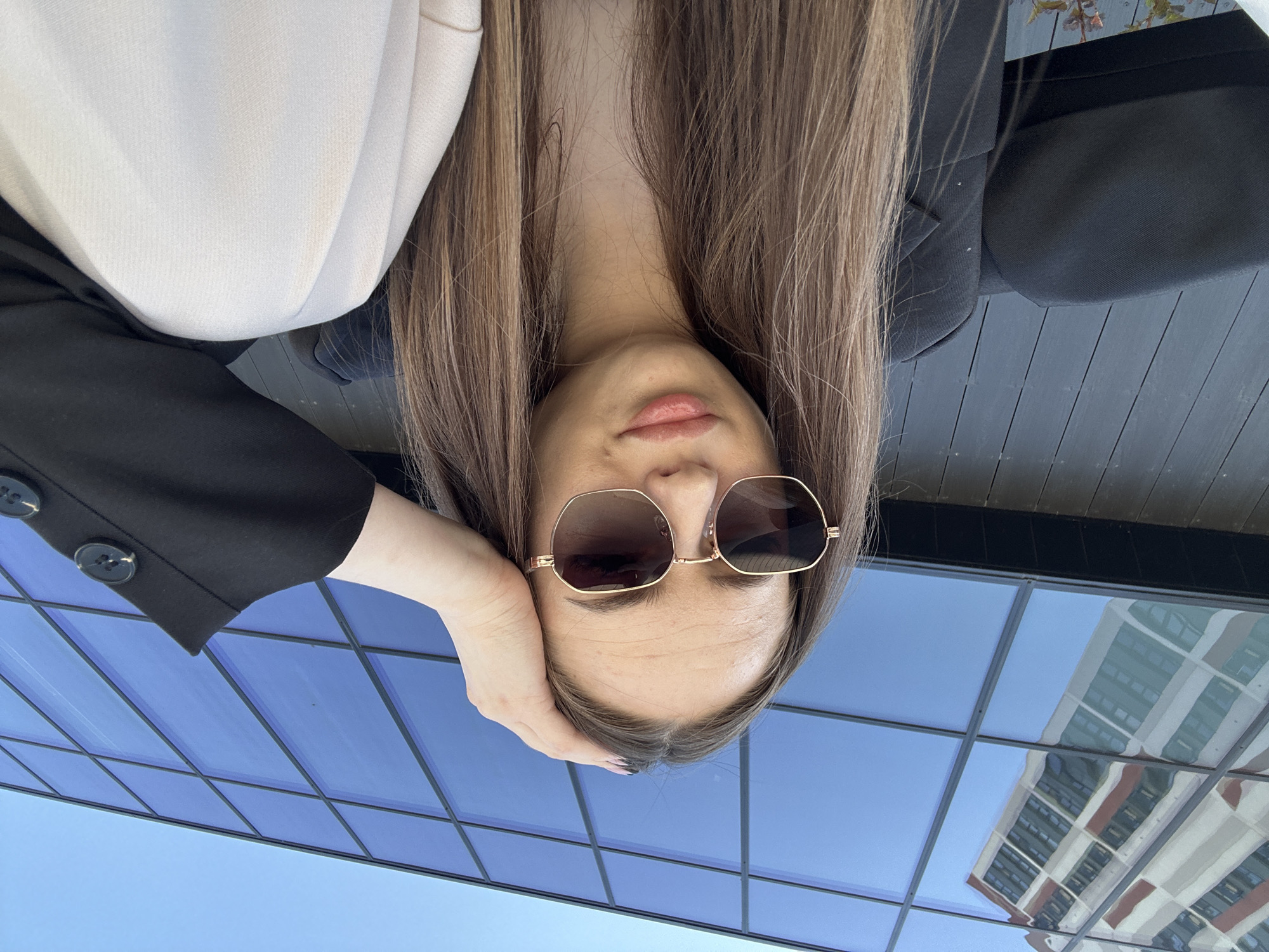 A young woman with long brown hair is facing the camera, wearing sunglasses, a light-colored shirt, and a dark jacket. She is posing with her head resting on her hand. The background features a modern building with blue-tinted windows, which reflect another building. The image is taken from a low angle. The city and location are unknown.
