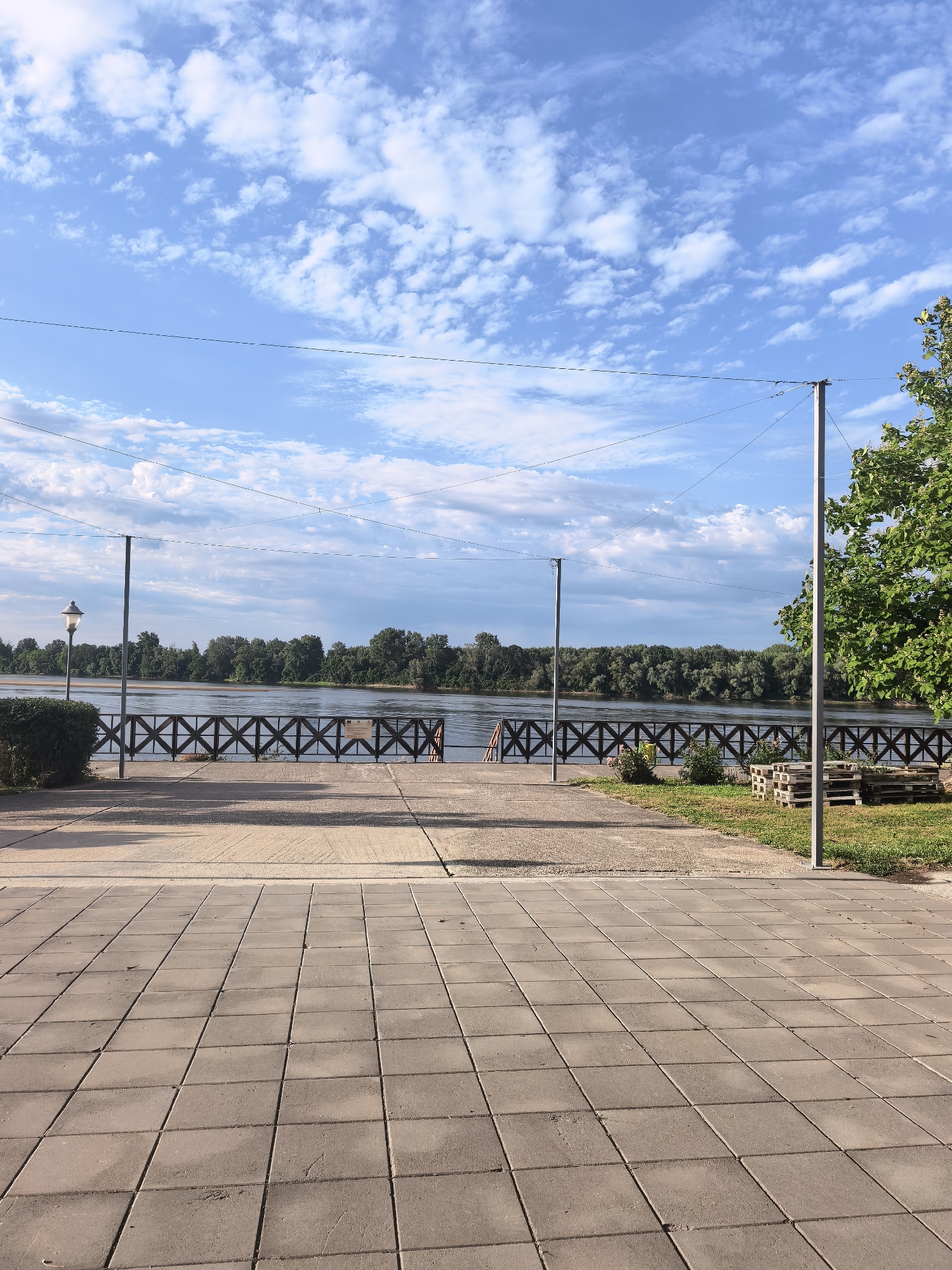 This is a photograph of the Danube River waterfront in Belene, Bulgaria, on a clear day. The foreground is dominated by a paved area made of square concrete tiles. Beyond this, there is a grassy area with some wooden pallets stacked. A decorative wooden fence runs along the edge of the promenade. The river is wide and calm, with a dense line of trees on the opposite bank. The sky is a bright blue with scattered white clouds. Two tall poles with wires stretched between them are visible in the middle ground, along with a lamppost to the left. The overall impression is one of a pleasant, well-maintained public space.