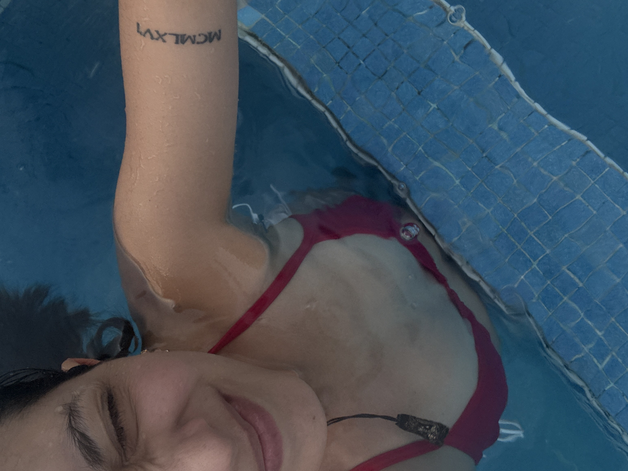 A woman with dark hair and closed eyes floats in a swimming pool with blue tiled edges. She is wearing a red bikini. A tattoo of "MCMLXVI" is visible on her left arm. The scene is in Veracruz City, Mexico.