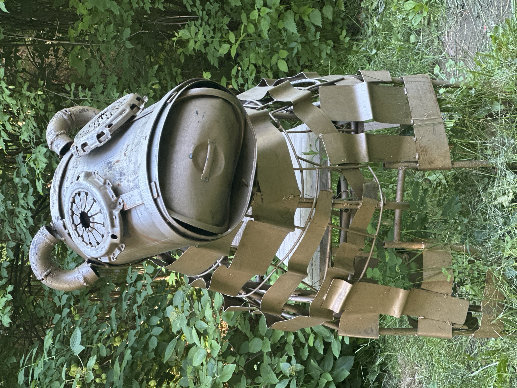 An outdoor, eye-level, full-shot photograph displays a metallic, bear-shaped sculpture situated amidst dense green foliage. The sculpture's head is constructed from a large metal canister with two curved pipes extending upwards, resembling ears. On the front of the head, a circular, fan-like component is visible on each side, and the snout is formed by a smaller, oval-shaped metal piece with a subtle indentation for a mouth. The body of the bear is a cage-like structure made of flat, bent metal pieces attached to a framework of pipes. The sculpture is painted a muted metallic brown and appears to be weathered. The background consists of an abundance of green leaves and branches, suggesting a natural, possibly wooded, environment. The ground beneath the sculpture is covered in grass and some sparse plants. There are no people present in the image.