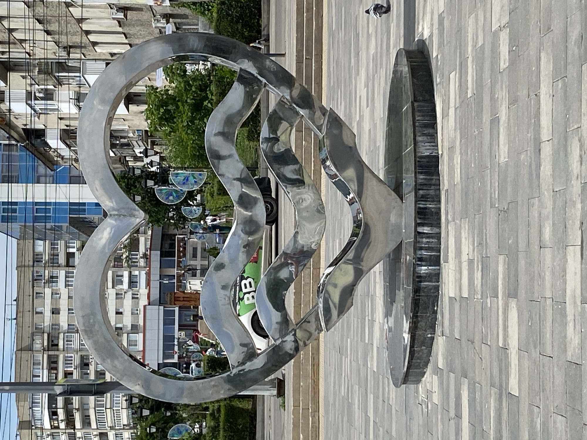 This is a vertically oriented, full-shot image of a modern metal sculpture in an urban outdoor setting. The sculpture is a series of interconnected, flowing metal shapes that appear to be made of polished chrome or stainless steel, reflecting the surrounding environment. A large heart shape forms the left side of the sculpture, seamlessly transitioning into three undulating wavy forms that lead to a circular base on the right. The sculpture is positioned on a light-gray tiled pavement, which forms a grid pattern.
In the background, a multi-story building with numerous windows is visible, suggesting a city environment. Trees with lush green foliage are also present, peeking out from behind the sculpture and the building. Several small, decorative glass orbs with intricate patterns are hanging from unseen supports, some of which are partially visible in the mid-ground. A white vehicle with green lettering is parked behind the sculpture.
A person is faintly visible in the distance, walking on the tiled area. Another person is also seen in the background on the left side. The lighting suggests it is daytime, likely sunny, as evidenced by the reflections on the sculpture. The overall scene conveys a sense of modern urban design and public art. The green lettering on the vehicle appears to say "BB", but the rest is illegible.