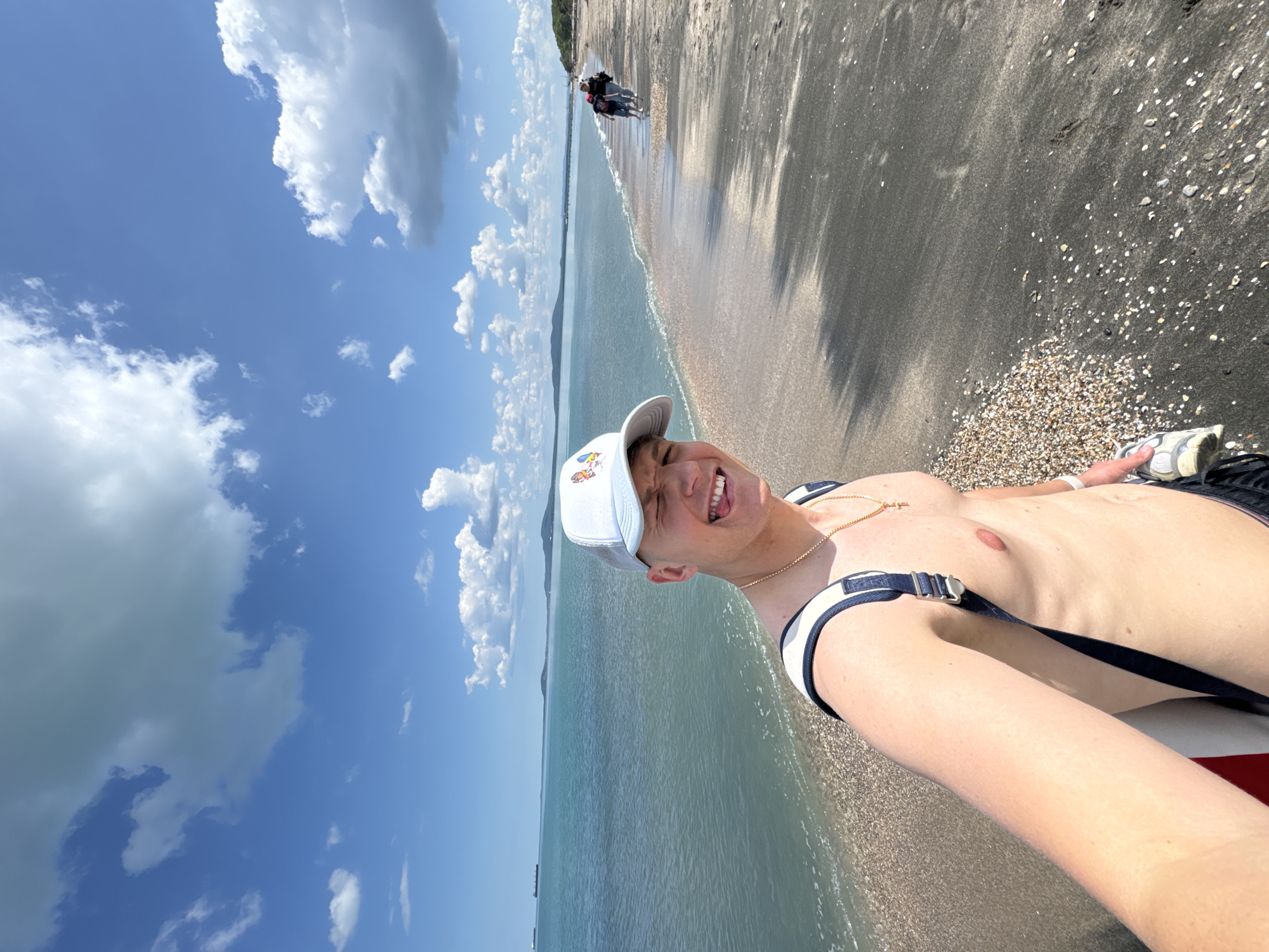 A fair-skinned young man is pictured in a selfie while on a beach in Burgas, Bulgaria. He wears a white baseball cap, a gold necklace with a cross, and is shirtless. He is standing on a beach with dark sand, with a shoreline in the background that stretches to the horizon. The sky is blue with scattered clouds. In the distance, two people can be seen walking along the shoreline.