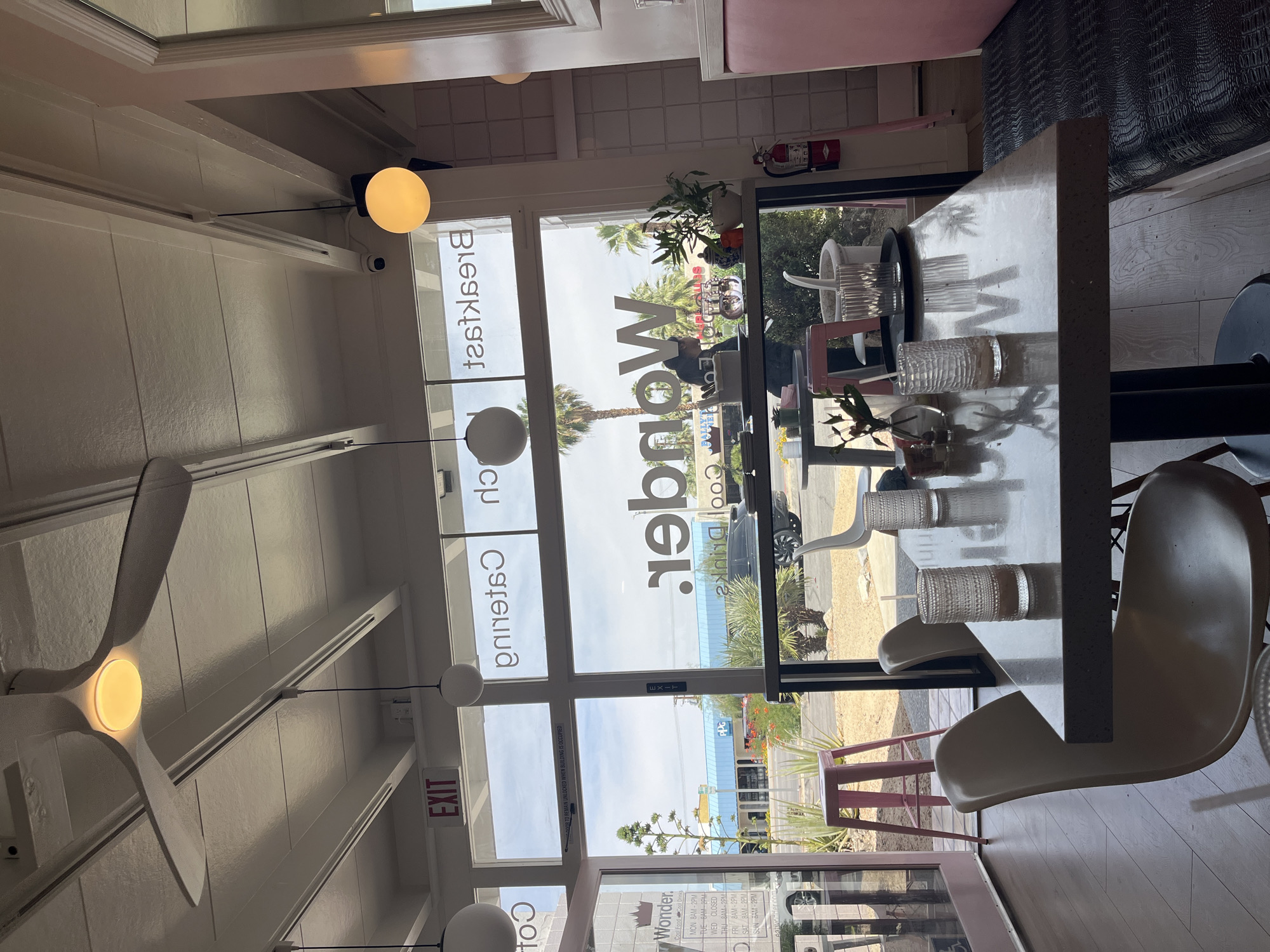 Here's a forensic description of the image:
**Scene:** Interior of a dining establishment, possibly a cafe or restaurant, located in Palm Desert, United States of America.
**Environment:** The space is brightly lit, appearing to have large windows that offer an exterior view. There is an exit sign visible.
**Objects:**
* Tables and chairs are present. The chairs are white in color with a distinctive modern design.
* Various objects (glassware, decorative items) are on the table.
* A ceiling fan is visible, with a yellow light.
* A fire extinguisher is mounted on the wall near the top of the window.
**Subjects:**
* The windows feature text indicating the name "Wonder," along with phrases like "Cool Drinks" and menu or business hours.
* Outside of the windows, there are palm trees, buildings, and cars visible, suggesting an urban or suburban location.
**Actions/Interactions:** No people are actively present or engaged in any visible action.