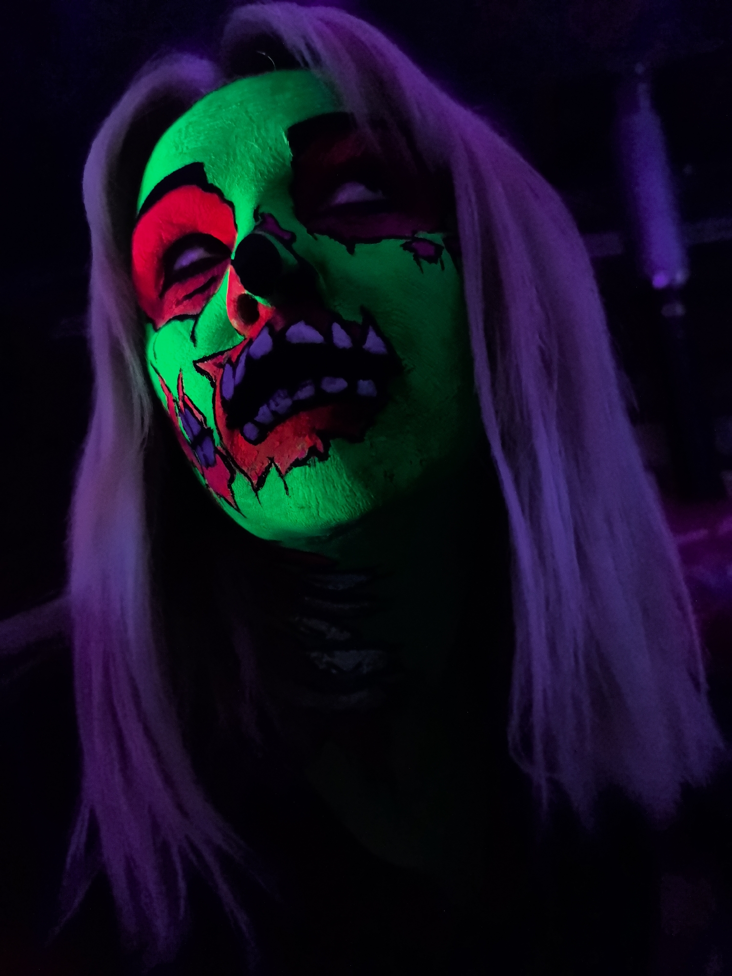 In a dimly lit environment, possibly a room or a club with blacklights, a person is depicted with dramatic neon green and red face paint. The makeup is designed to resemble a zombie or a skull, with exaggerated features such as empty eye sockets filled with red, a jagged mouth with black and purple teeth, and sharp red and purple accents on the cheeks and forehead, all illuminated by the fluorescent glow. The person has long, light-colored hair, appearing to be either blonde or bleached, which is tinged with purple light. The background is dark and indistinct, with faint purple light sources visible on the right side, suggesting an artificial lighting setup. The image appears to be a close-up portrait, focusing entirely on the subject's face and the vibrant, unsettling makeup. There is no identifiable information about the specific location within Noviy, Russia.