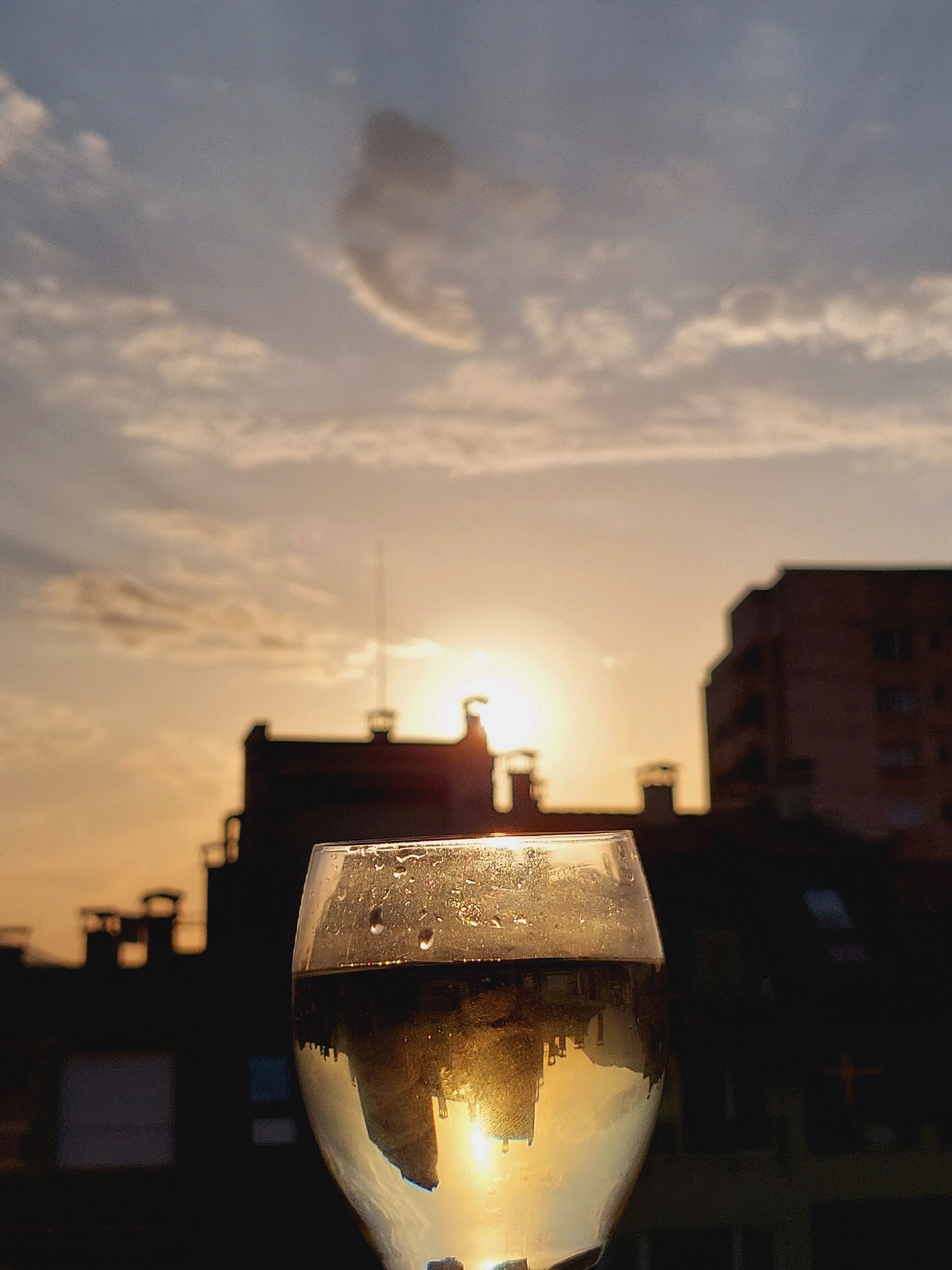 A wine glass is positioned in the foreground, filled with a light-colored liquid, likely white wine. The glass has water droplets on its exterior, indicating it is chilled. The liquid inside reflects the setting sun and the silhouettes of buildings. The background shows a cityscape at dusk, with the sun partially obscured by clouds and buildings. The sky is a warm gradient of orange and yellow, transitioning to blue higher up. The buildings are dark silhouettes against the bright sky, suggesting they are residential or office buildings. The overall scene is tranquil, evoking a sense of peace and relaxation as the day ends. The presence of buildings with chimneys and antennas indicates an urban setting. This image was taken in Sofia, Bulgaria.