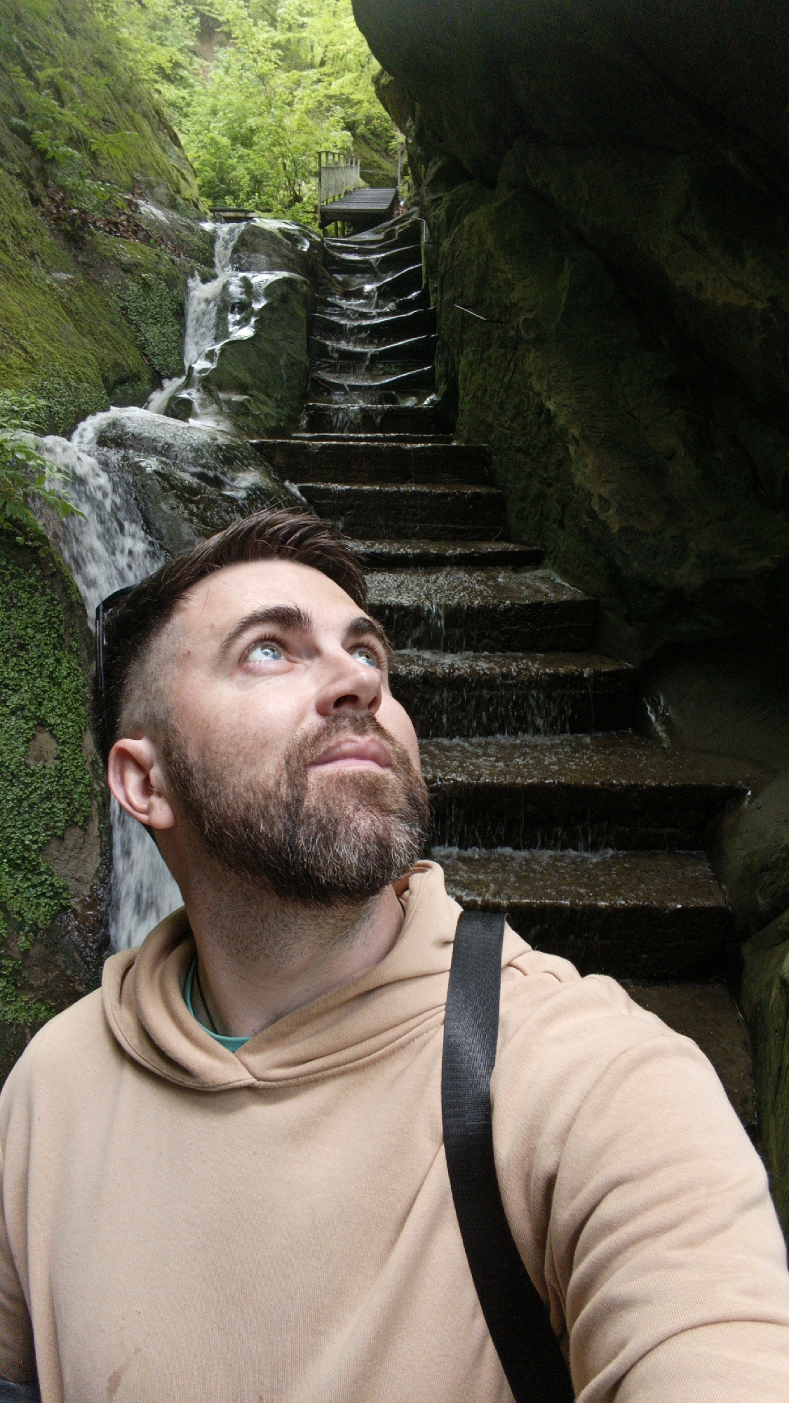 A man with blue eyes and a beard stands in front of a stone staircase with a waterfall running down it, located within a mossy, rocky environment. He wears a tan hoodie and a black strap crosses his body. The location is likely in or near the city of Goryachiy Klyuch, Russia. A wooden bridge is visible above the waterfall at the top of the steps. The man is looking upwards.